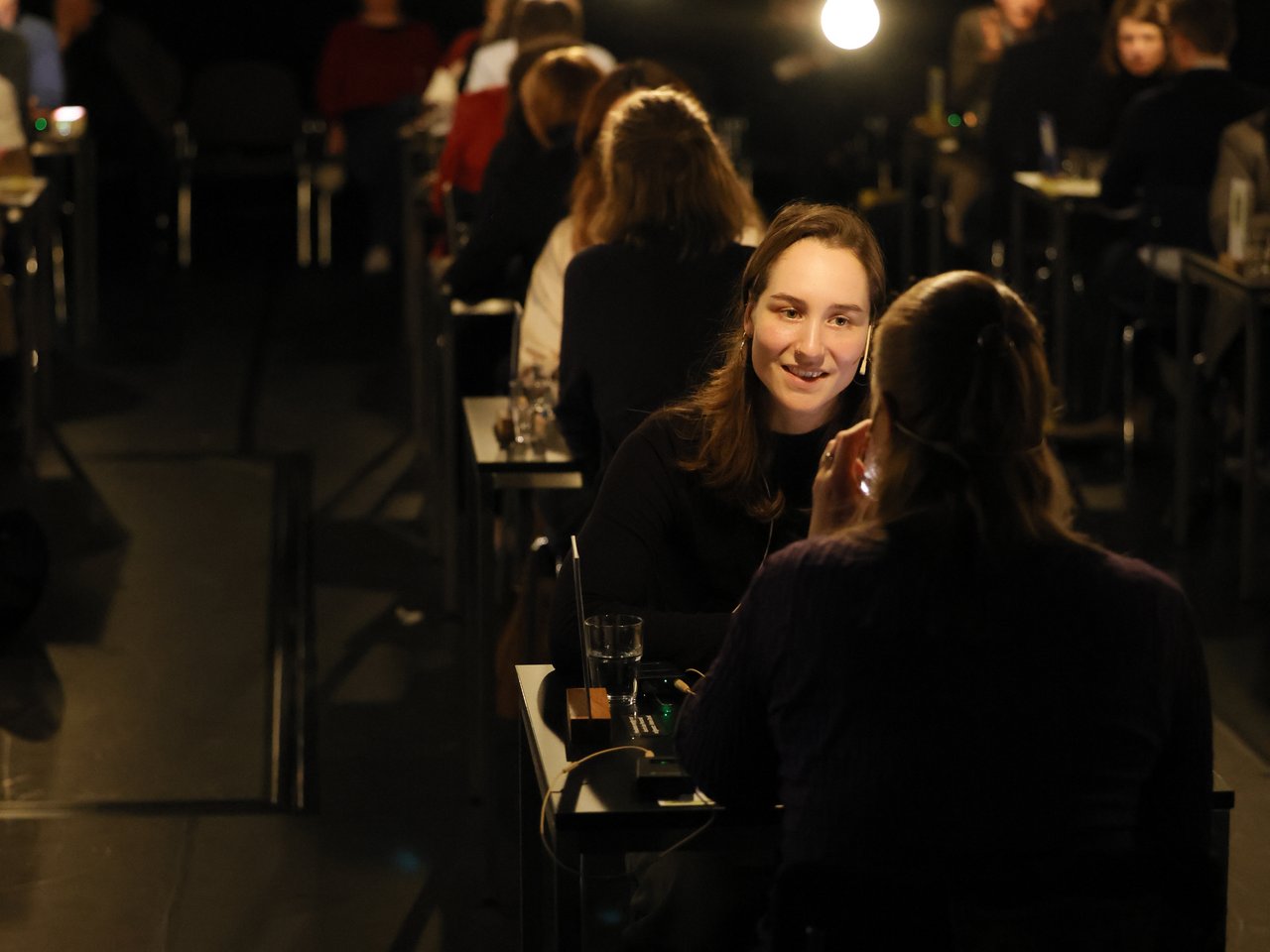Zwei Frauen sitzen sich beim Markt für nützliches Wissen und Nicht-Wissen im neuen theater in Halle (Saale) an einem kleinen Tisch gegenüber und unterhalten sich. Eine der Frauen lächelt ihr Gegenüber an. Auf dem Tisch stehen ein Wasserglas und ein kleines Audiogerät. Im Hintergrund finden zahlreiche weitere Einzelgespräche statt. Hängende Glühbirnen beleuchten den ansonsten dunklen Veranstaltungsraum.