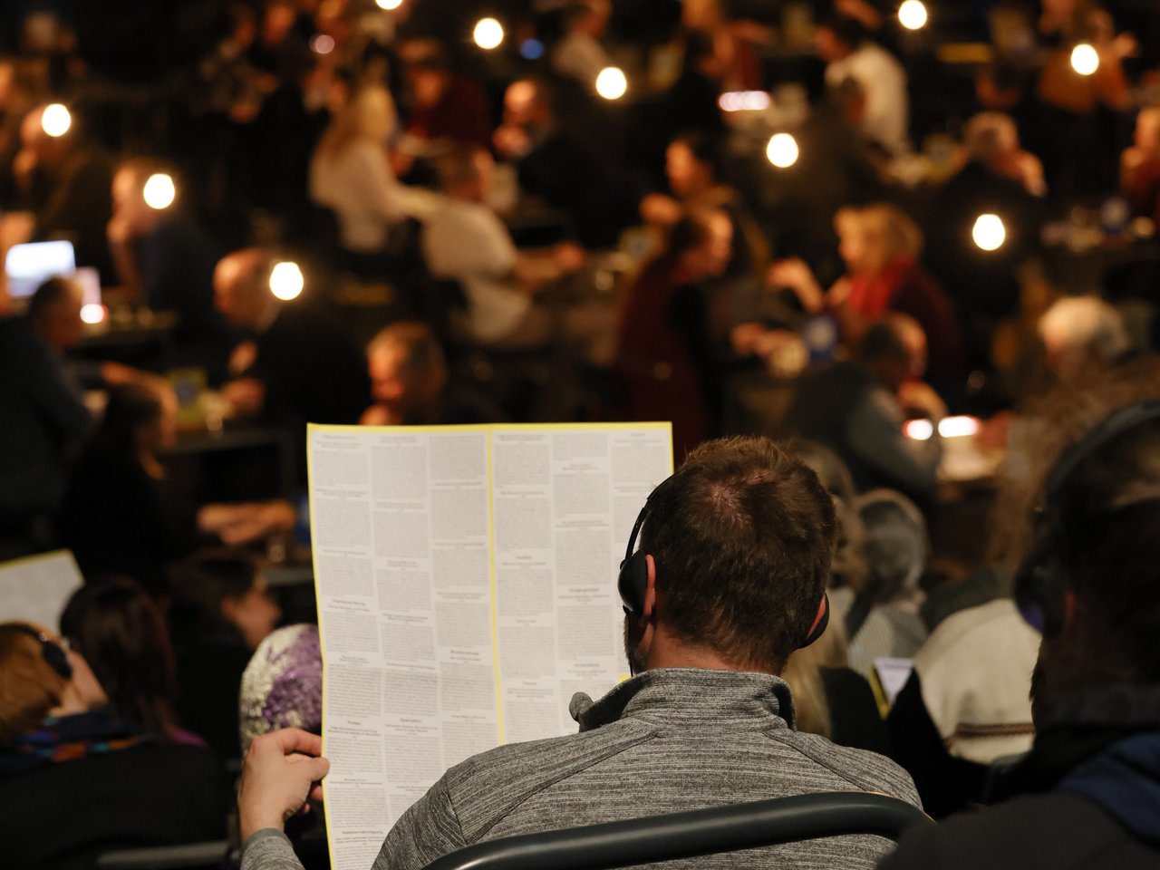 Ein Besucher mit Kopfhörern sitzt auf den ansteigenden Zuschauerreihen beim Markt für nützliches Wissen und Nicht-Wissen im neuen theater in Halle (Saale). Er hält die aufgefaltete Enzyklopädie in den Händen und blickt auf die Gesprächsarena. Im unscharfen Hintergrund sitzen zahlreiche Menschen an kleinen Tischen und tauschen sich aus. Hängende Glühbirnen beleuchten den abgedunkelten Raum.