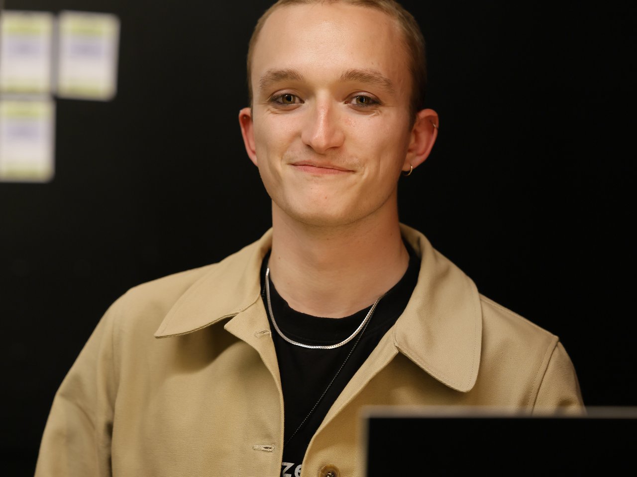 Ein junger Mitarbeiter mit kurzen Haaren, beigefarbener Jacke und schwarzem T-Shirt sitzt lächelnd hinter dem Check-in-Tresen beim Markt für nützliches Wissen und Nicht-Wissen im neuen theater in Halle (Saale). Vor ihm liegen Informationskarten auf dem grünen Tresen. Rechts steht ein schwarzes Schild mit der Aufschrift „AUSVERKAUFT“.