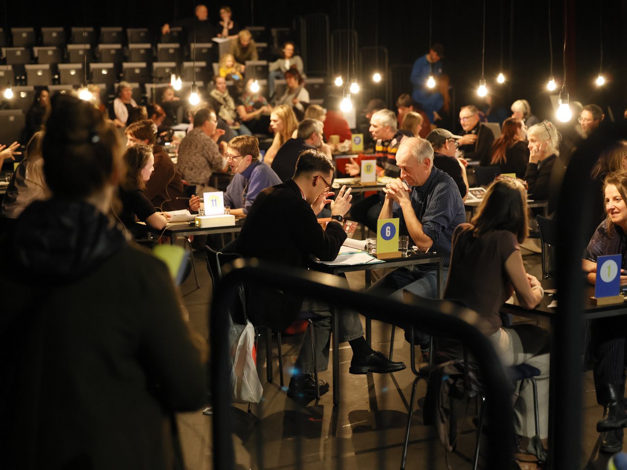 Blick in die gut besuchte Arena beim Markt für nützliches Wissen und Nicht-Wissen im neuen theater in Halle (Saale). An zahlreichen nummerierten Tischen sitzen sich Expert:innen und Besucher:innen gegenüber und tauschen Wissen und Erfahrungen aus. Einige gestikulieren während der Gespräche, andere hören aufmerksam zu. Im Hintergrund sitzen weitere Gäste auf den ansteigenden Zuschauerreihen. Zahlreiche hängende Glühbirnen beleuchten den abgedunkelten Veranstaltungsraum.