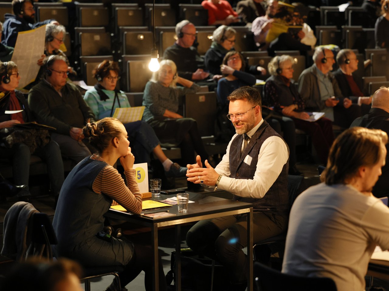 Ein Bestatter mit Brille, Bart und dunkler Weste sitzt beim Markt für nützliches Wissen und Nicht-Wissen im neuen theater in Halle (Saale) an Tisch Nummer 5. Er spricht mit einer Frau, die ihm aufmerksam zuhört, und gestikuliert dabei mit beiden Händen. Im Hintergrund sitzen zahlreiche Besucher:innen mit Kopfhörern auf den ansteigenden Zuschauerreihen und verfolgen die Gespräche in der Arena.