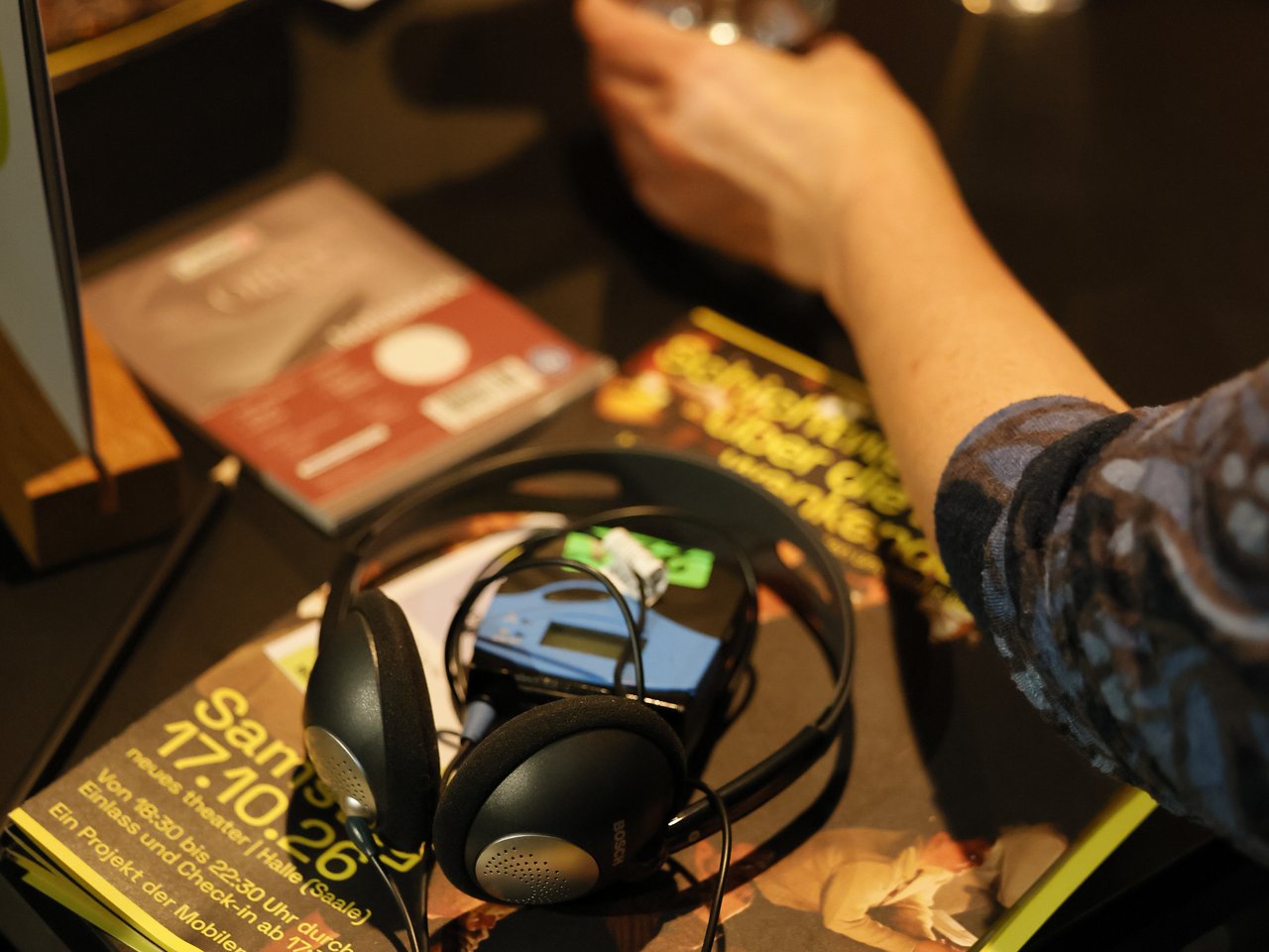Detailaufnahme eines Gesprächstisches beim Markt für nützliches Wissen und Nicht-Wissen im neuen theater in Halle (Saale). Auf dem Tisch liegen die Enzyklopädie, Informationsmaterialien, ein Kopfhörer und ein kleines Audiogerät. Im Hintergrund gestikuliert eine Person während eines Gesprächs, während eine andere ein Wasserglas in der Hand hält.
