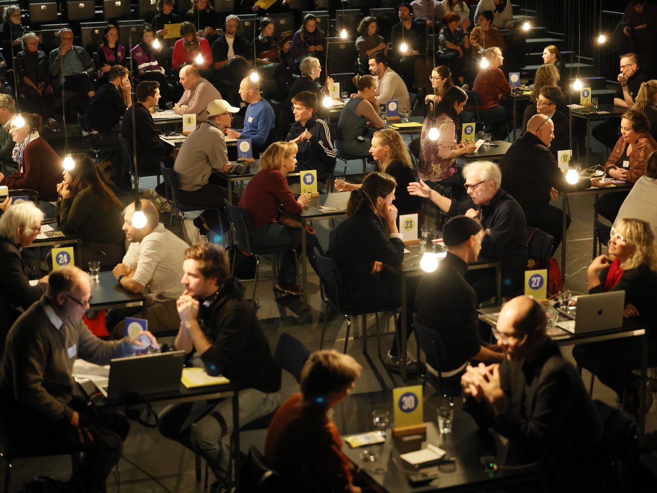 Blick von oben auf die gut besuchte Arena beim Markt für nützliches Wissen und Nicht-Wissen im neuen theater in Halle (Saale). An zahlreichen nummerierten Tischen sitzen sich Expert:innen und Besucher:innen gegenüber und tauschen Wissen und Erfahrungen aus. Einige gestikulieren lebhaft, andere hören aufmerksam zu. Auf den ansteigenden Zuschauerreihen verfolgen weitere Gäste die Gespräche, teilweise über Kopfhörer. Hängende Glühbirnen beleuchten die Arena.