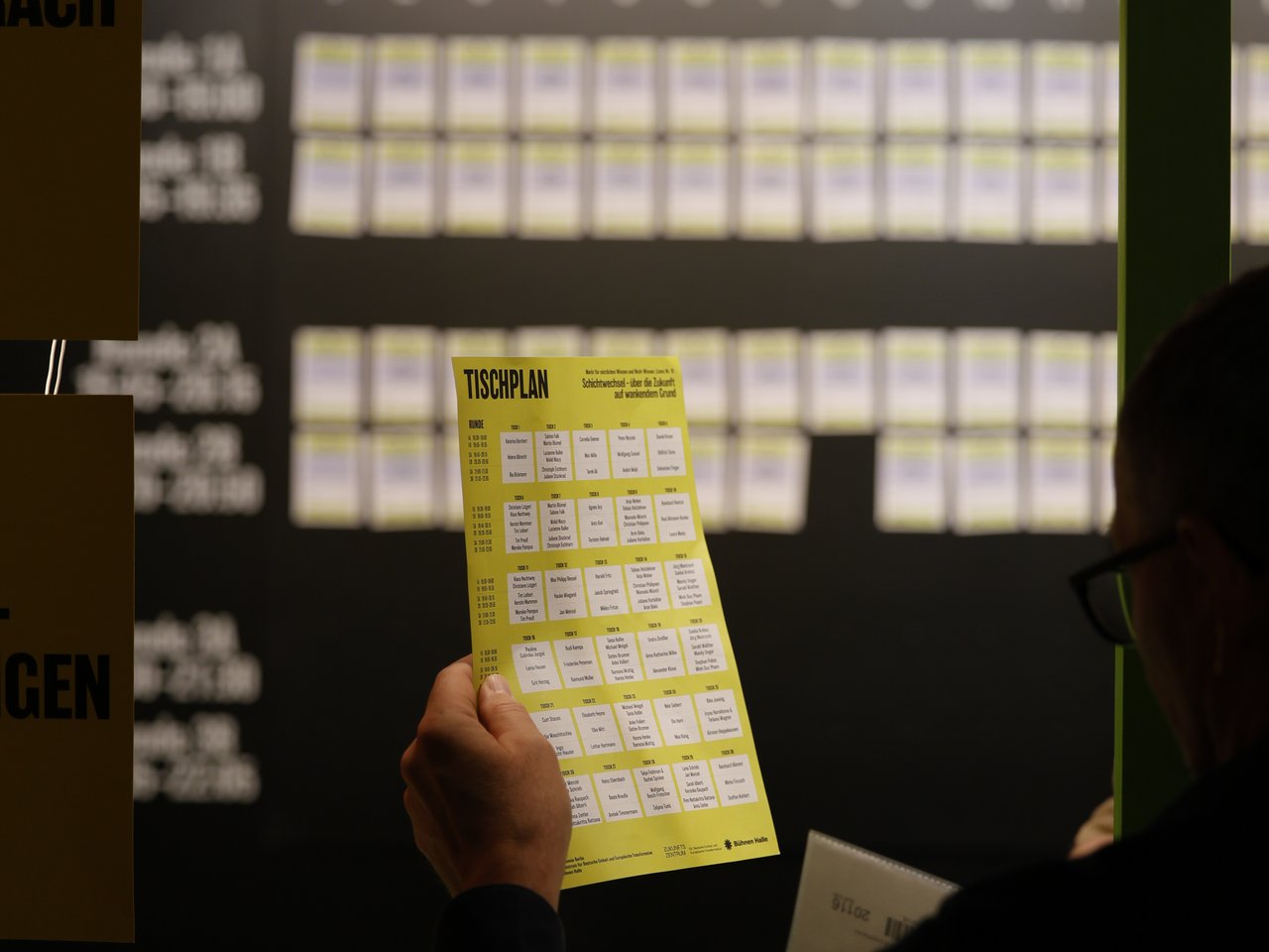 Ein Besucher mit Brille betrachtet am Check-in-Tresen beim Markt für nützliches Wissen und Nicht-Wissen im neuen theater in Halle (Saale) einen gelben Tischplan. Darauf sind die Gesprächsangebote und ihre jeweiligen Tischnummern verzeichnet. Im unscharfen Hintergrund hängt eine große Übersicht mit den nummerierten Gesprächstischen und den verschiedenen Gesprächsrunden.