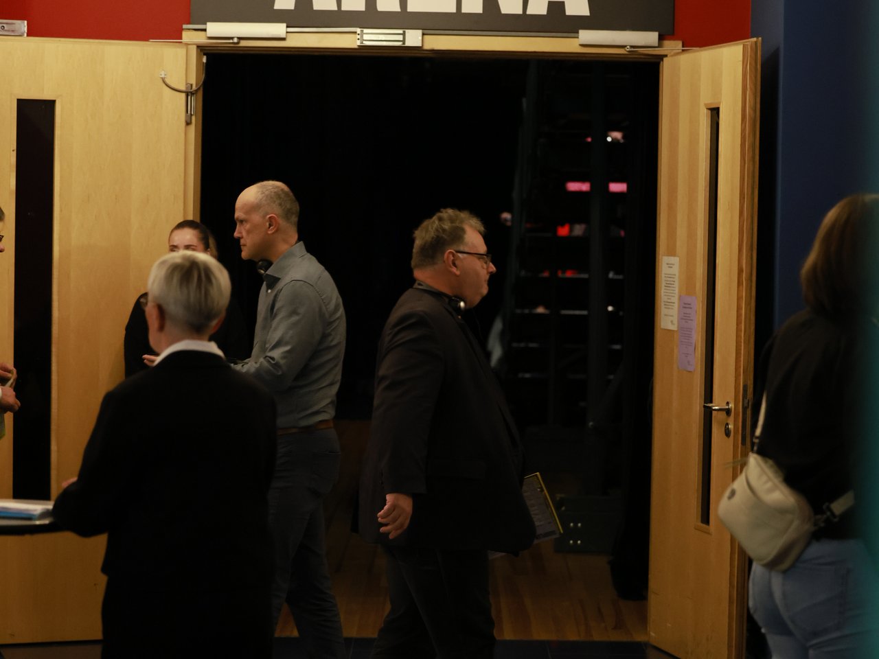 Mehrere Besucher:innen stehen im Eingangsbereich der Arena beim Markt für nützliches Wissen und Nicht-Wissen im neuen theater in Halle (Saale). Über den geöffneten Flügeltüren hängt ein großes schwarzes Schild mit der weißen Aufschrift „ARENA“. Die Wände sind leuchtend rot gestrichen, an der Decke hängt ein Kronleuchter.