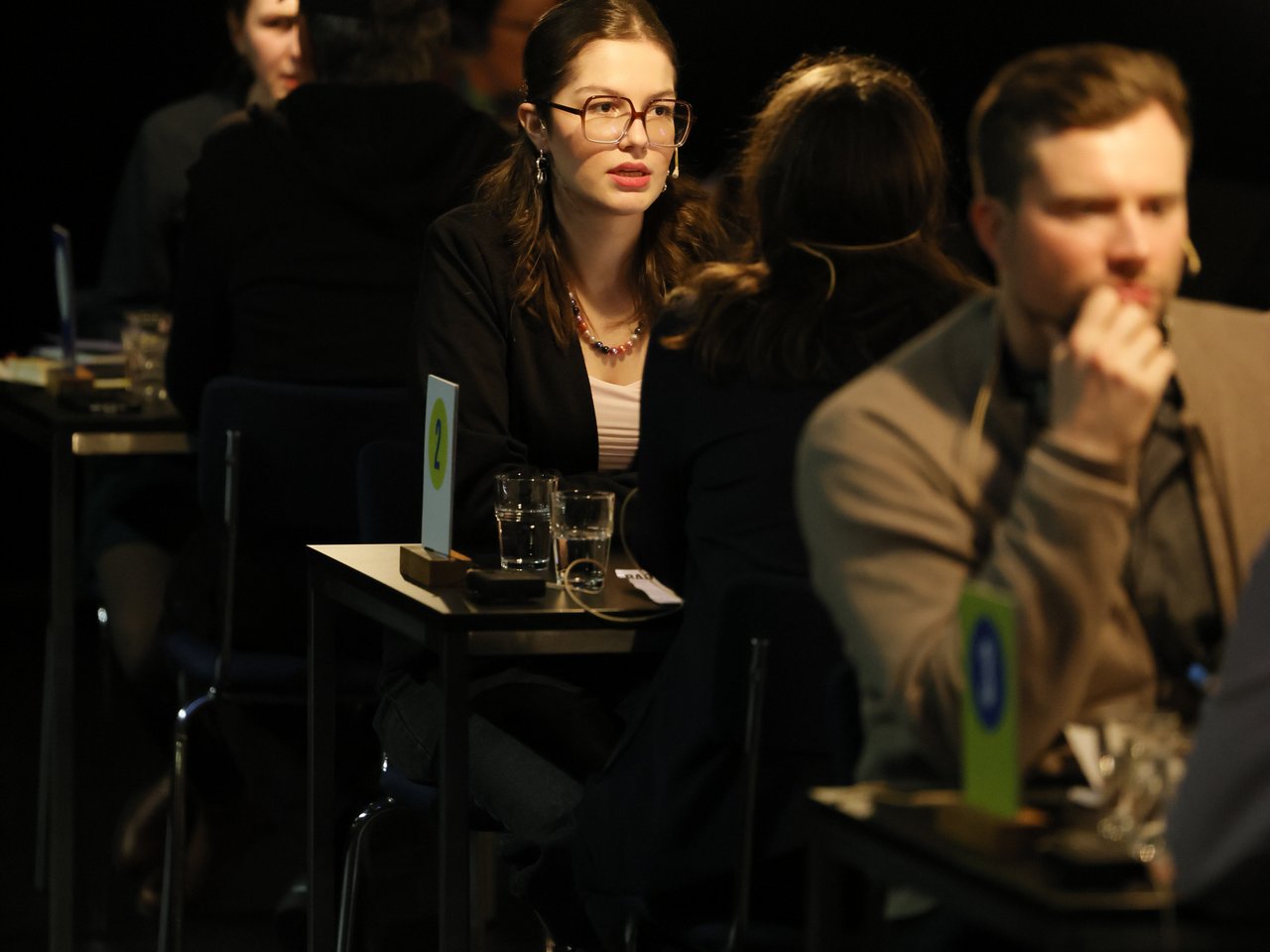 Eine junge Frau mit langen braunen Haaren, Brille und schwarzem Blazer sitzt beim Markt für nützliches Wissen und Nicht-Wissen im neuen theater in Halle (Saale) an Tisch Nummer 2. Sie spricht mit einer Frau, die ihr den Rücken zuwendet. Im Vordergrund ist ein weiterer Gesprächsteilnehmer zu sehen. Hängende Glühbirnen beleuchten die einzelnen Gesprächsrunden im abgedunkelten Veranstaltungsraum.