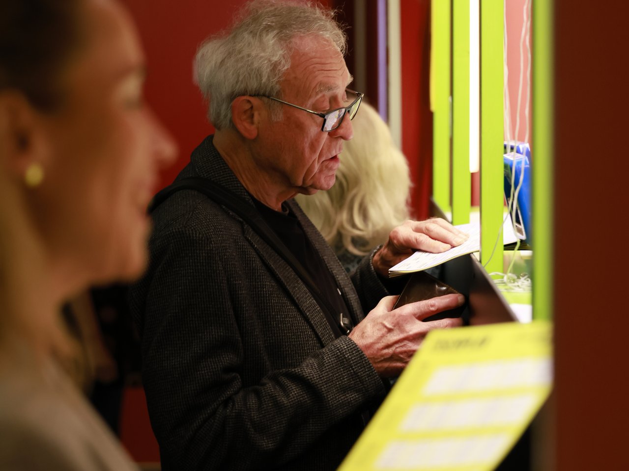 Ein älterer Mann mit grauen Haaren und Brille steht am Check-in-Tresen beim Markt für nützliches Wissen und Nicht-Wissen im neuen theater in Halle (Saale). Er betrachtet eine Karte und hält eine Geldbörse in der Hand. Im unscharfen Vordergrund liest eine Frau die Enzyklopädie..