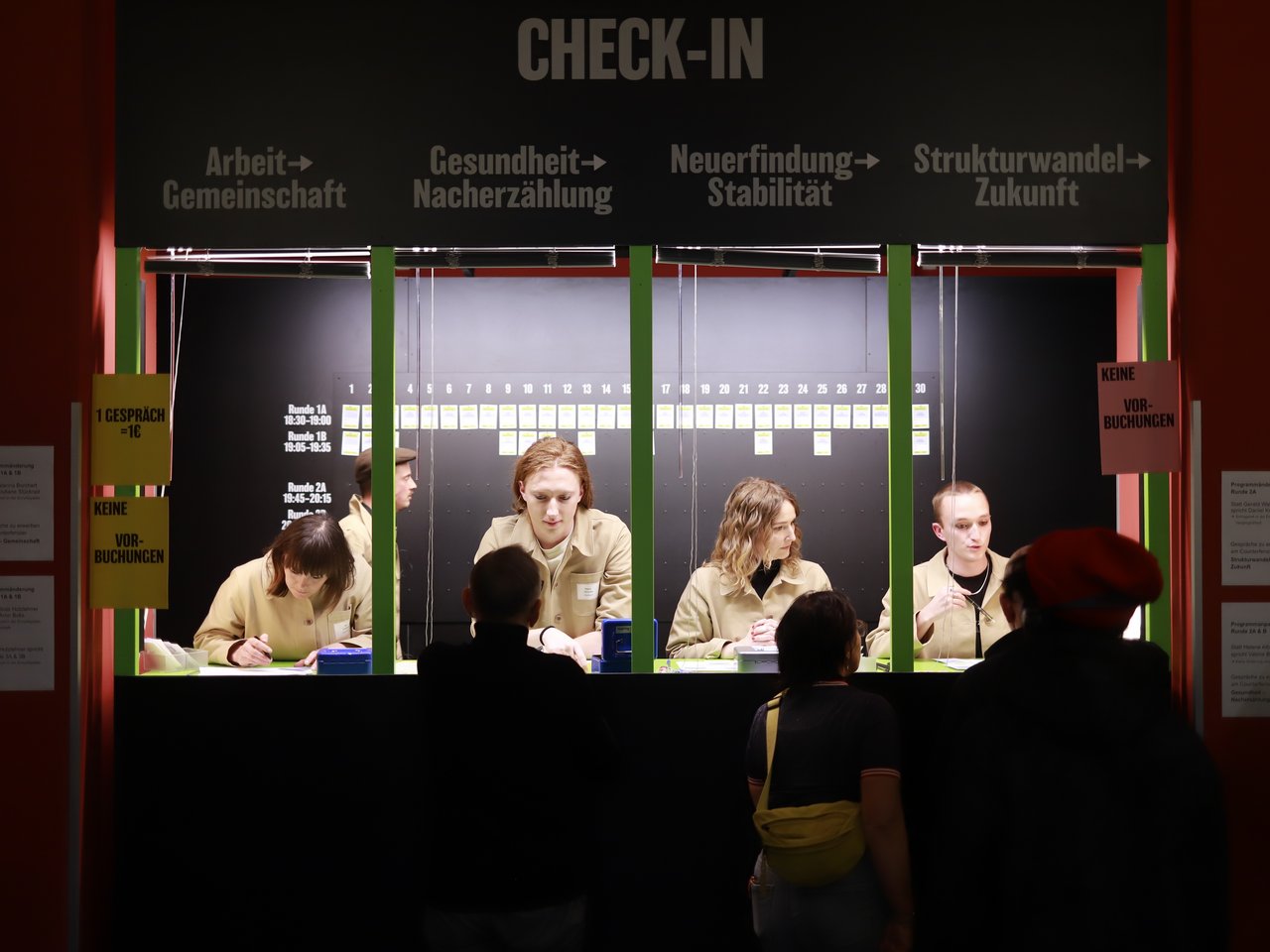 Blick auf den Check-in-Bereich beim Markt für nützliches Wissen und Nicht-Wissen im neuen theater in Halle (Saale). Vier Mitarbeitende sitzen hinter einem langen Tresen und empfangen die ankommenden Besucher:innen. Über dem Tresen steht „CHECK-IN“, darunter sind vier Themenbereiche ausgeschildert: Arbeit, Gesundheit, Neuerfindung und Strukturwandel. Im Vordergrund warten mehrere Gäste.