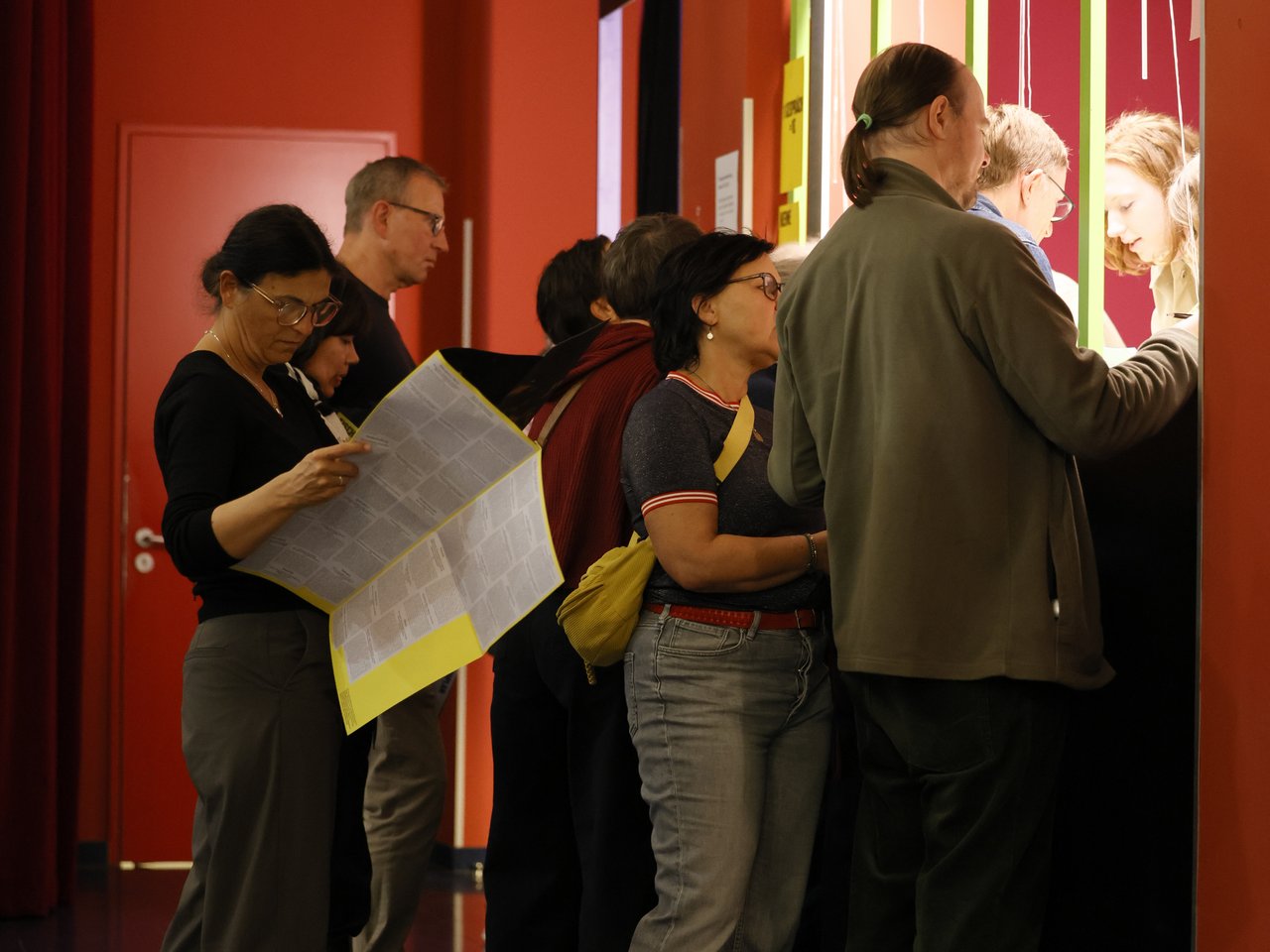 Mehrere Besucher:innen stehen am Check-in-Tresen beim Markt für nützliches Wissen und Nicht-Wissen im neuen theater in Halle (Saale). Eine Frau im Vordergrund studiert die großformatige Enzyklopädie, während andere Gäste am Tresen mit den Mitarbeitenden sprechen. Die roten Wände und grünen Elemente des Check-in-Bereichs prägen die Gestaltung.