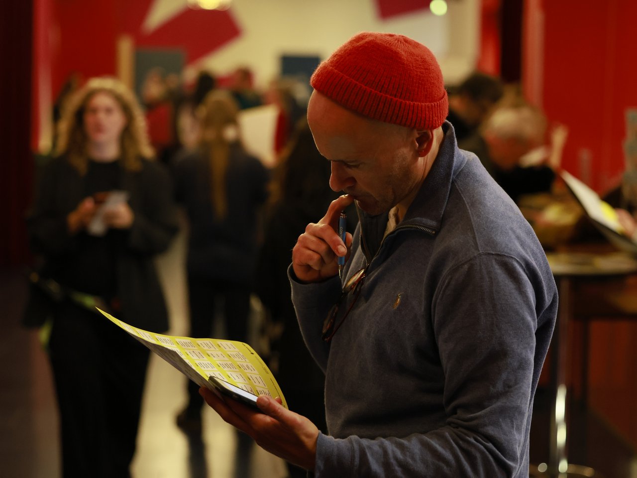Ein Mann mit roter Strickmütze und blauem Pullover betrachtet konzentriert die Enzyklopädie beim Markt für nützliches Wissen und Nicht-Wissen im neuen theater in Halle (Saale). Er hält einen Stift an seine Lippen. Im unscharfen Hintergrund stehen weitere Besucher:innen im rot gestalteten Foyer