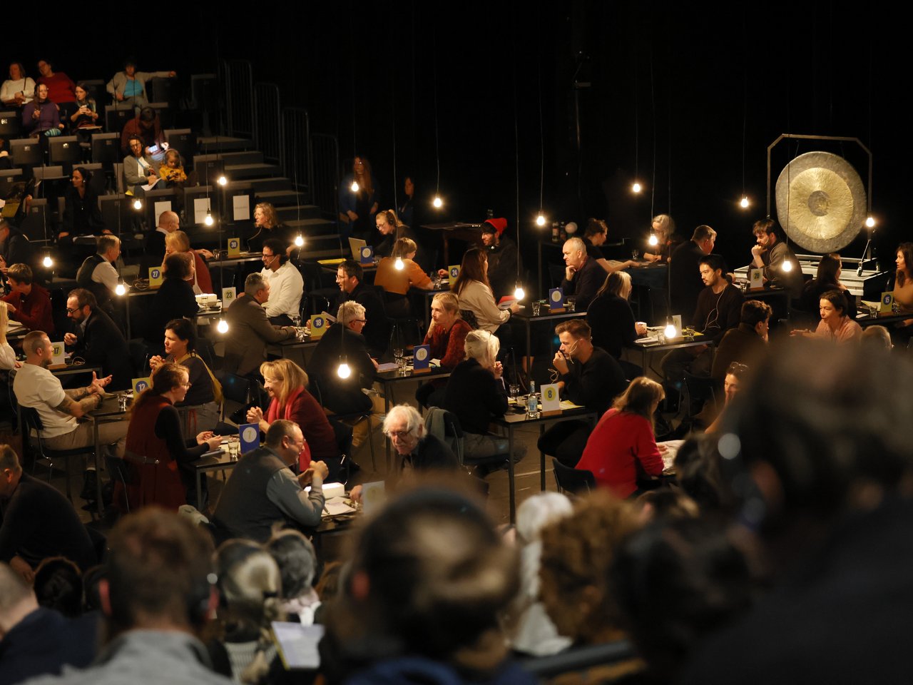 Blick von oben auf die gut besuchte Gesprächsarena beim Markt für nützliches Wissen und Nicht-Wissen im neuen theater in Halle (Saale). An zahlreichen nummerierten Tischen sitzen sich jeweils zwei Personen gegenüber und tauschen Wissen und Erfahrungen aus. Hängende Glühbirnen beleuchten die einzelnen Gesprächsrunden im abgedunkelten Raum. Auf den ansteigenden Sitzreihen verfolgen weitere Besucher:innen das Geschehen. Im Hintergrund steht ein großer goldfarbener Gong.
