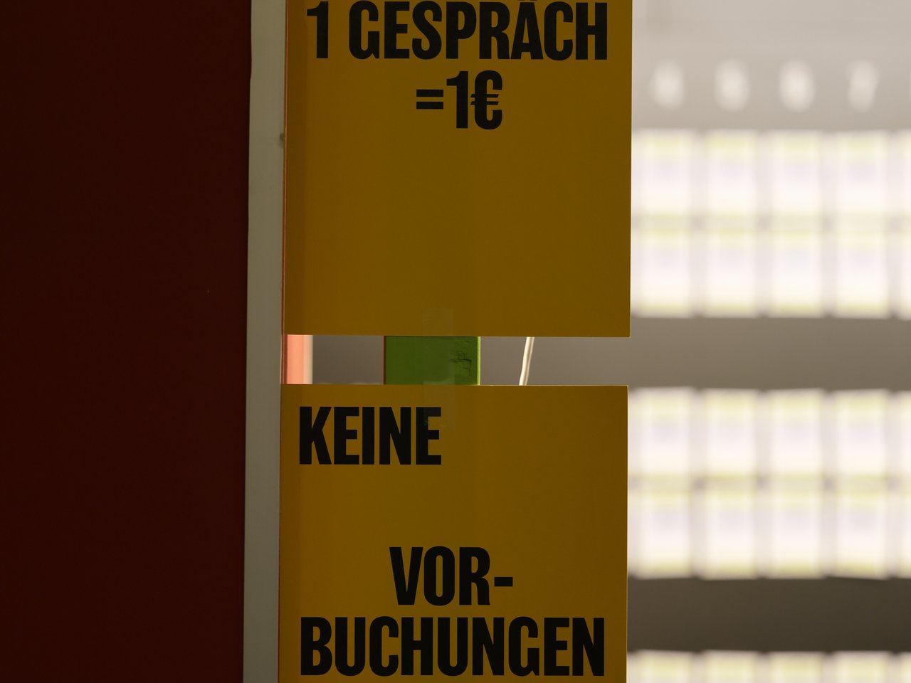 Zwei gelbe Hinweisschilder am Check-in-Tresen beim Markt für nützliches Wissen und Nicht-Wissen im neuen theater in Halle (Saale). Auf den Schildern stehen in großen schwarzen Buchstaben die Hinweise „1 Gespräch = 1 €“ und „KEINE VORBUCHUNGEN“. Im unscharfen Hintergrund ist die Übersicht der Gesprächsrunden zu sehen.