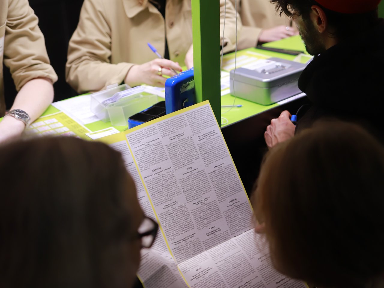 Mehrere Besucher:innen stehen am Check-in-Tresen beim Markt für nützliches Wissen und Nicht-Wissen im neuen theater in Halle (Saale). Eine Besucherin hält die aufgefaltete Enzyklopädie in den Händen, während eine Mitarbeiterin hinter dem grün gestalteten Tresen Unterlagen ausfüllt. Neben ihr sitzt ein weiterer Mitarbeiter. Ein Besucher mit roter Mütze wartet am Tresen.