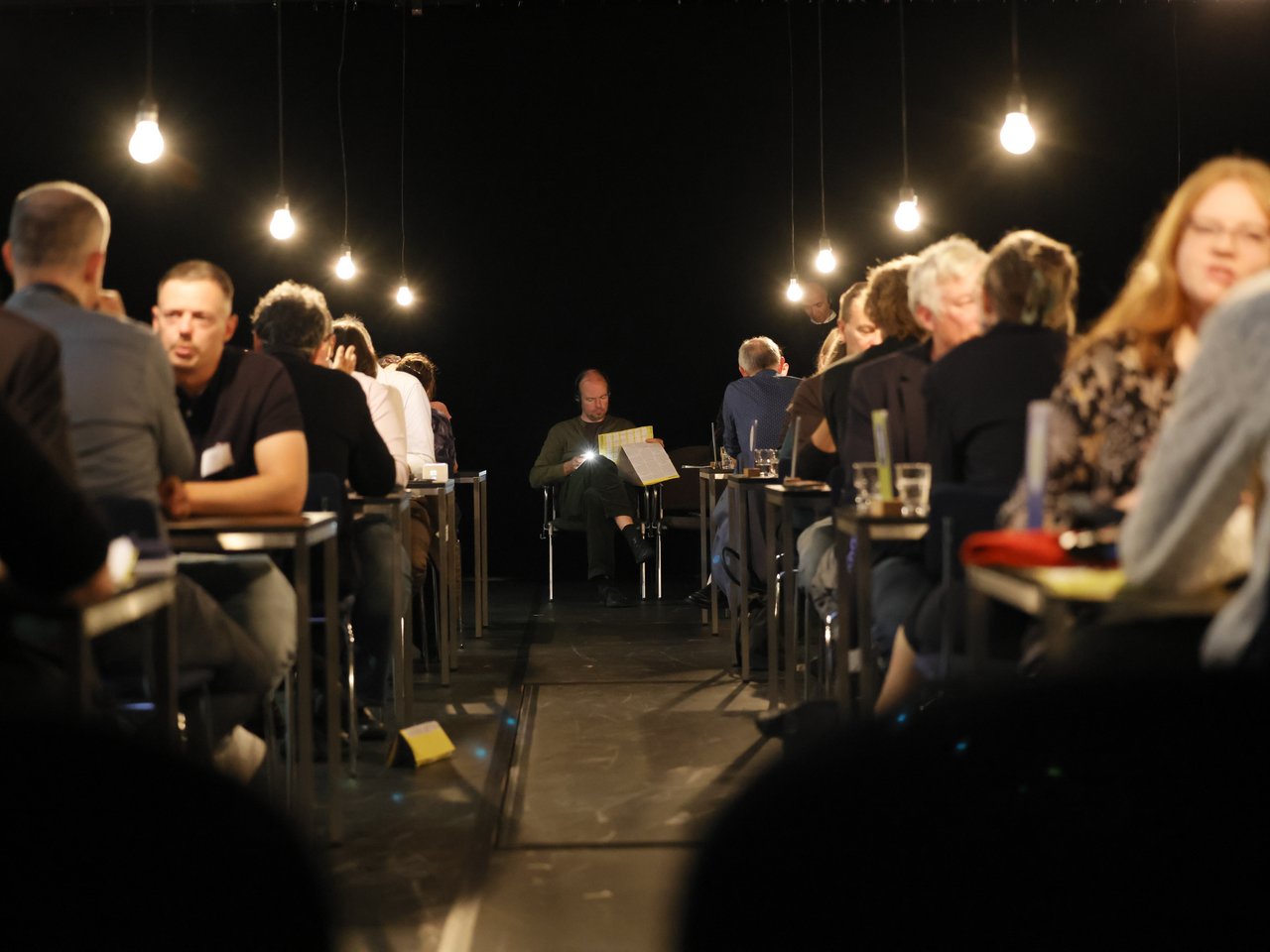 Blick durch einen Mittelgang in die Arena beim Markt für nützliches Wissen und Nicht-Wissen im neuen theater in Halle (Saale). Links und rechts sitzen sich zahlreiche Expert:innen und Besucher:innen an kleinen Tischen gegenüber und führen Gespräche. Am Ende des Gangs sitzt ein Mann allein auf einem Stuhl und liest in der Enzyklopädie. Hängende Glühbirnen beleuchten den ansonsten dunklen Raum.