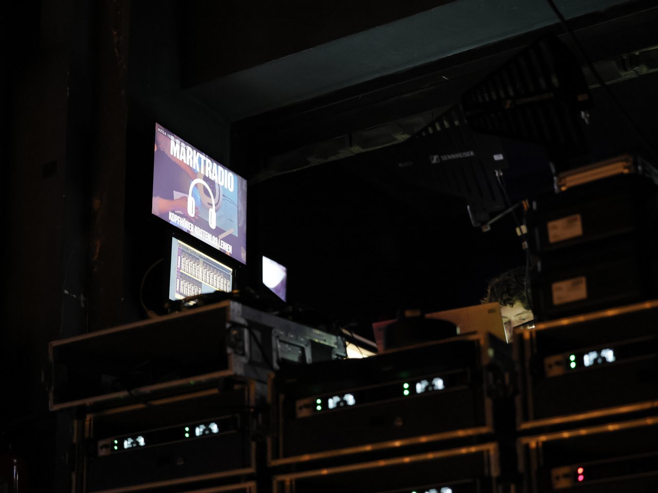 Blick auf die Technik des Marktradios beim Markt für nützliches Wissen und Nicht-Wissen im neuen theater in Halle (Saale). In einem abgedunkelten Technikbereich stehen mehrere Racks mit Audioequipment, Funkempfängern und Monitoren. Auf einem Bildschirm ist der Schriftzug „MARKTRADIO – KOPFHÖRER KOSTENLOS LEIHEN“ zu sehen. Hinter den Geräten ist eine Person zu erkennen, die die Technik betreut.  Bildtitel:
