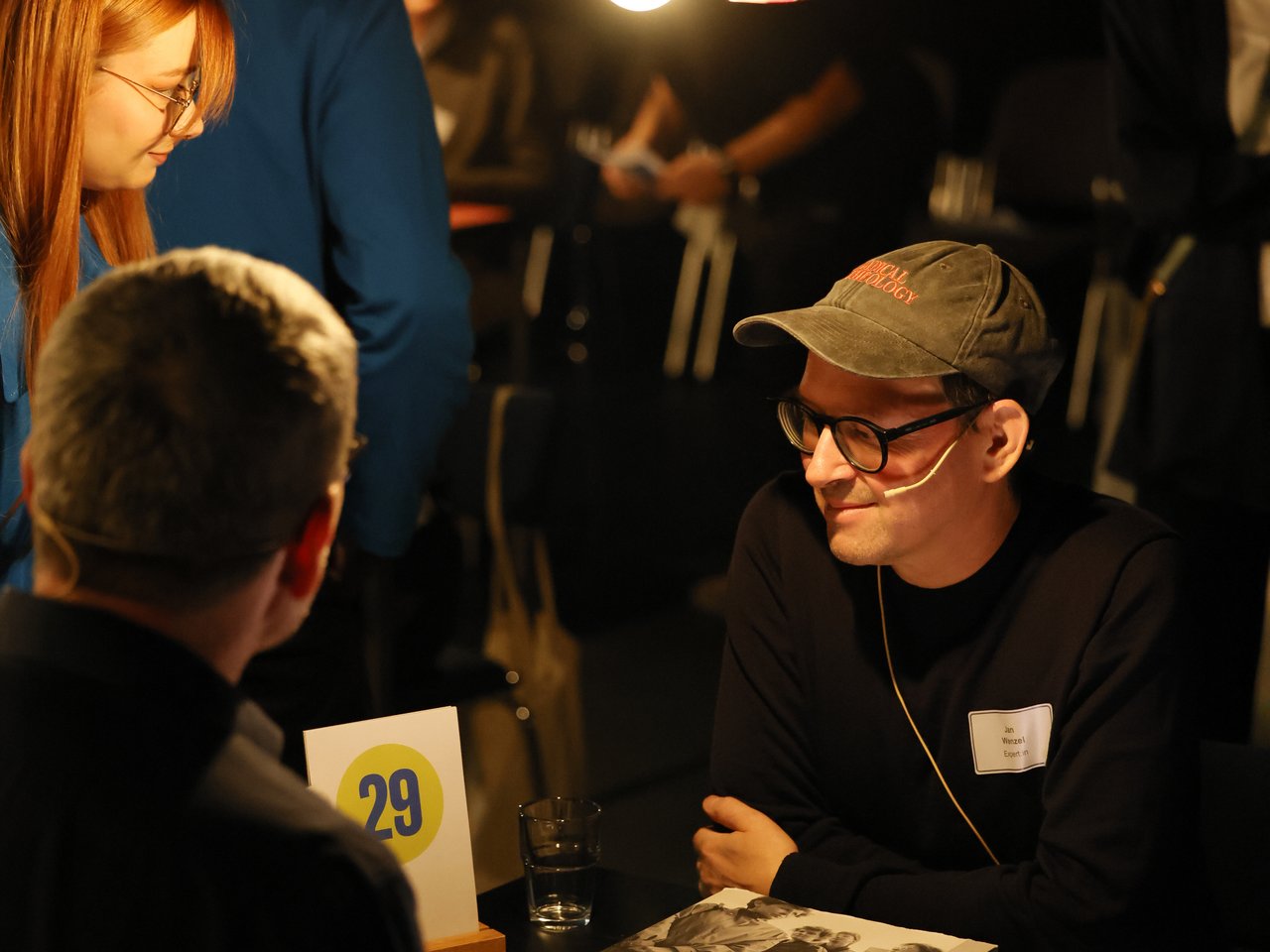 Zwei Männer sitzen sich beim Markt für nützliches Wissen und Nicht-Wissen im neuen theater in Halle (Saale) an Tisch Nummer 29 gegenüber und unterhalten sich. Einer der Männer trägt eine graue Baseballkappe, eine Brille und ein Headset. Auf dem Tisch liegen ein großformatiges Buch mit historischen Schwarz-Weiß-Fotografien, ein Wasserglas und weitere Materialien. Im Hintergrund sind weitere Besucher:innen und Veranstaltungspersonal zu sehen. Eine hängende Glühbirne beleuchtet die Gesprächssituation.