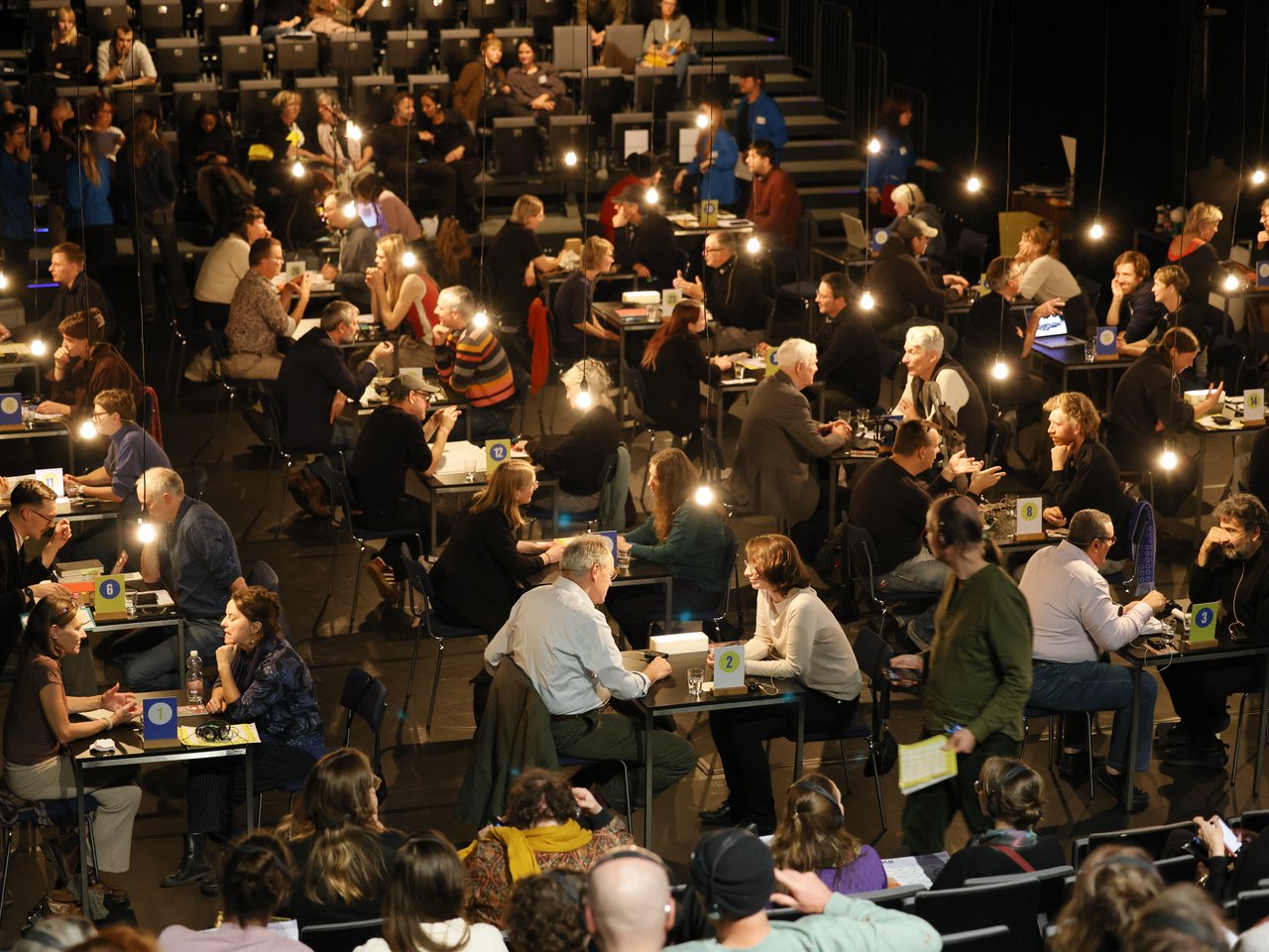 Blick von oben auf die gut besuchte Arena beim Markt für nützliches Wissen und Nicht-Wissen im neuen theater in Halle (Saale). An zahlreichen nummerierten Tischen sitzen sich Expert:innen und Besucher:innen gegenüber und tauschen Wissen und Erfahrungen aus. Einige gestikulieren lebhaft, andere hören aufmerksam zu. Auf den ansteigenden Zuschauerreihen verfolgen weitere Gäste die Gespräche, teilweise über Kopfhörer. Zahlreiche hängende Glühbirnen beleuchten den abgedunkelten Veranstaltungsraum.