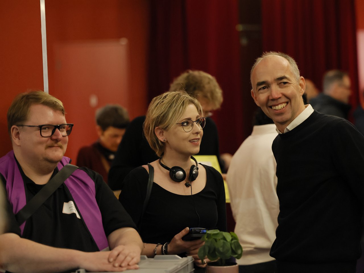 Holger Lemme, kaufmännischer Geschäftsführer des Zukunftszentrums, steht lächelnd neben zwei Personen beim Markt für nützliches Wissen und Nicht-Wissen im neuen theater in Halle (Saale). Links steht ein Mitarbeiter des Awareness-Teams mit lilafarbener Weste und Brille. Zwischen ihnen steht eine Frau mit kurzen blonden Haaren, Brille und Kopfhörern um den Hals. Im Hintergrund sind weitere Veranstaltungsgäste zu sehen.