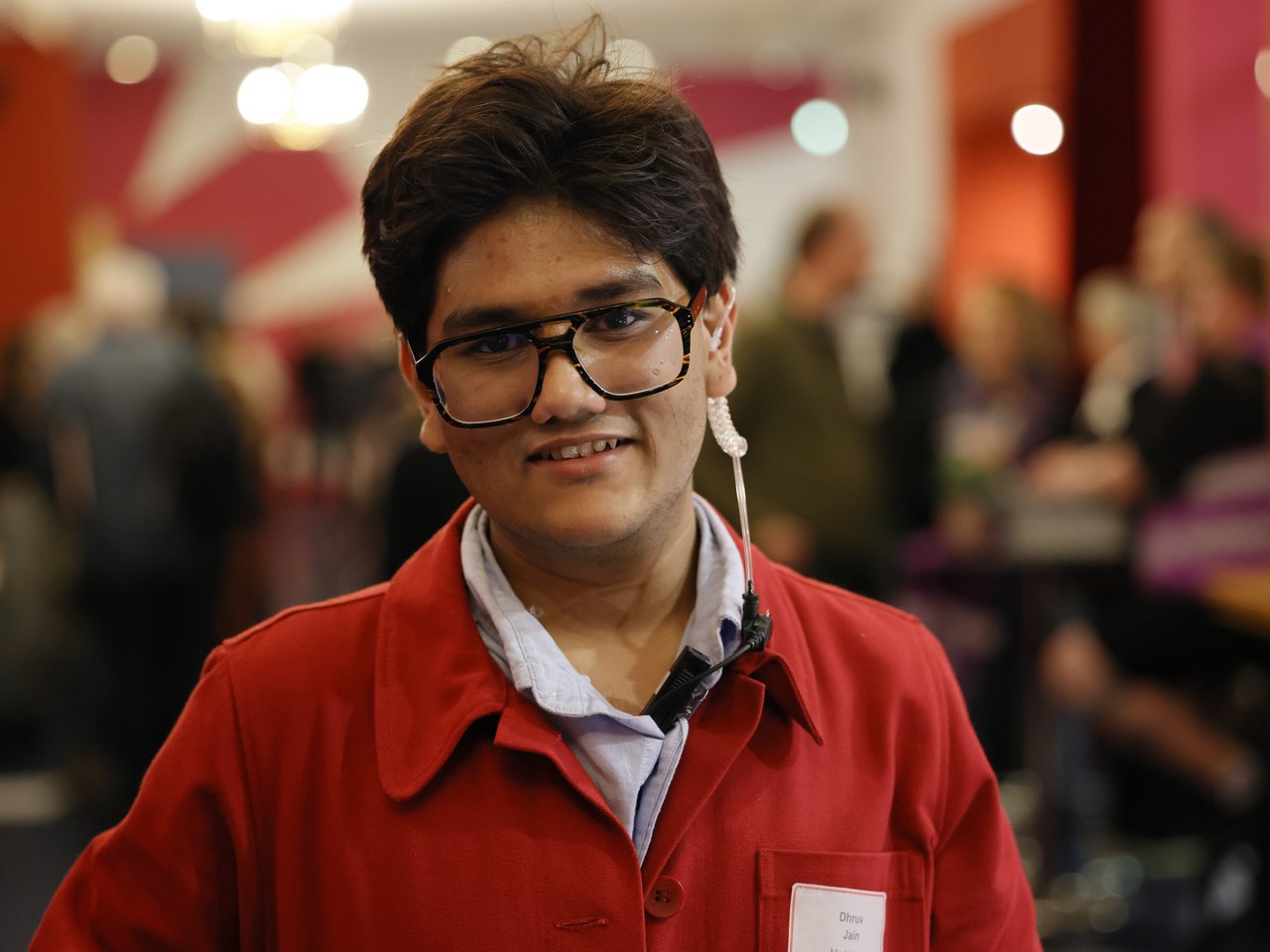 Dhruv Jain, Mitarbeiter des Marktteams beim Markt für nützliches Wissen und Nicht-Wissen im neuen theater in Halle (Saale), blickt lächelnd in die Kamera. Er trägt eine rote Jacke über einem hellblauen Hemd, eine schwarze Brille und einen Ohrhörer mit Spiralkabel. Im unscharfen Hintergrund sind zahlreiche Besucher:innen im Foyer zu sehen.
