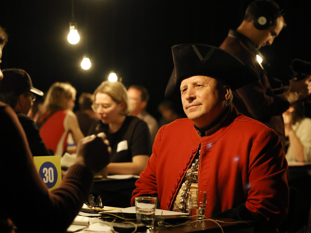 Ein Mann in historischer Kleidung mit rotem Gehrock, schwarzem Dreispitz und reich verzierter Weste sitzt beim Markt für nützliches Wissen und Nicht-Wissen im neuen theater in Halle (Saale) an Tisch Nummer 30. Er hört seinem Gegenüber aufmerksam zu. Auf dem Tisch stehen Wassergläser und liegen Gesprächsunterlagen. Im Hintergrund finden weitere Einzelgespräche statt. Hängende Glühbirnen beleuchten die abgedunkelte Arena.