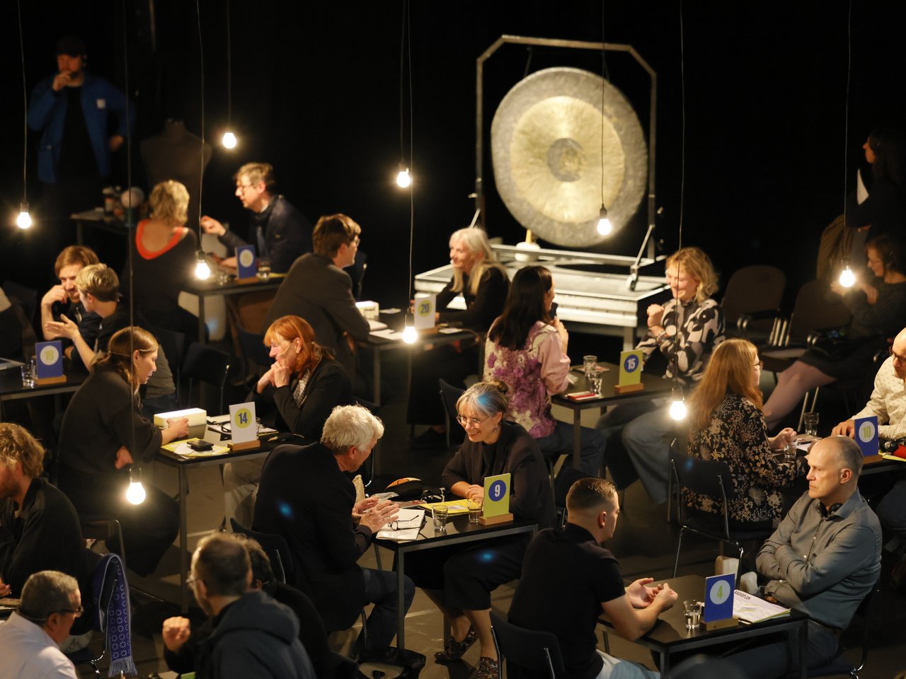 Blick von oben auf die Arena beim Markt für nützliches Wissen und Nicht-Wissen im neuen theater in Halle (Saale). An mehreren nummerierten Tischen sitzen sich Expert:innen und Besucher:innen gegenüber und tauschen Wissen und Erfahrungen aus. Einige gestikulieren, andere hören aufmerksam zu. Im Hintergrund steht ein großer goldfarbener Gong, der den Wechsel der Gesprächsrunden signalisiert. Hängende Glühbirnen beleuchten den abgedunkelten Raum.