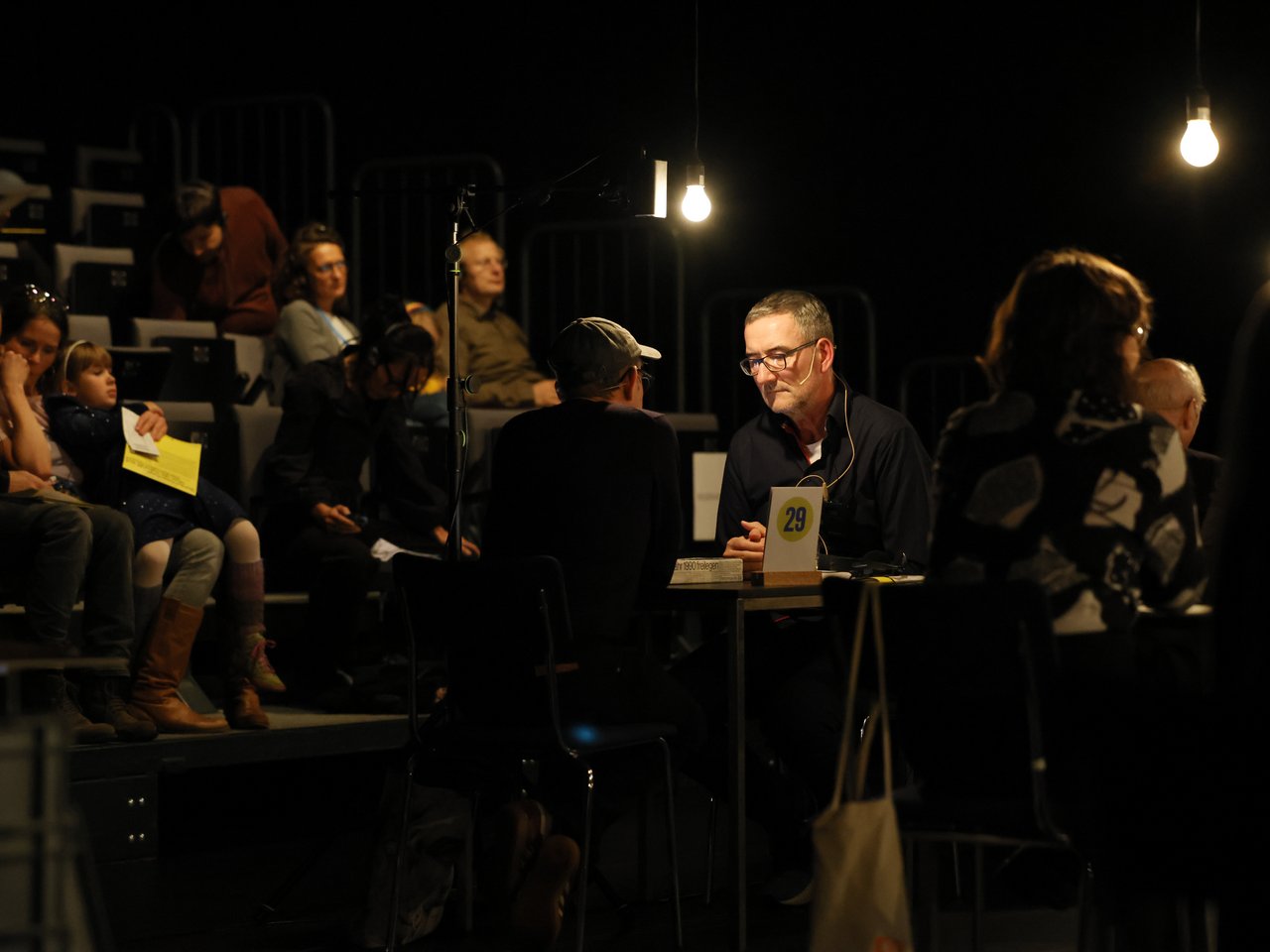 Zwei Männer sitzen sich beim Markt für nützliches Wissen und Nicht-Wissen im neuen theater in Halle (Saale) an Tisch Nummer 29 gegenüber und unterhalten sich. Einer der Männer trägt eine dunkle Baseballkappe, sein Gegenüber eine Brille und ein schwarzes Hemd. Beide tragen Headsets. Auf den ansteigenden Sitzreihen im Hintergrund sitzen weitere Besucher:innen.