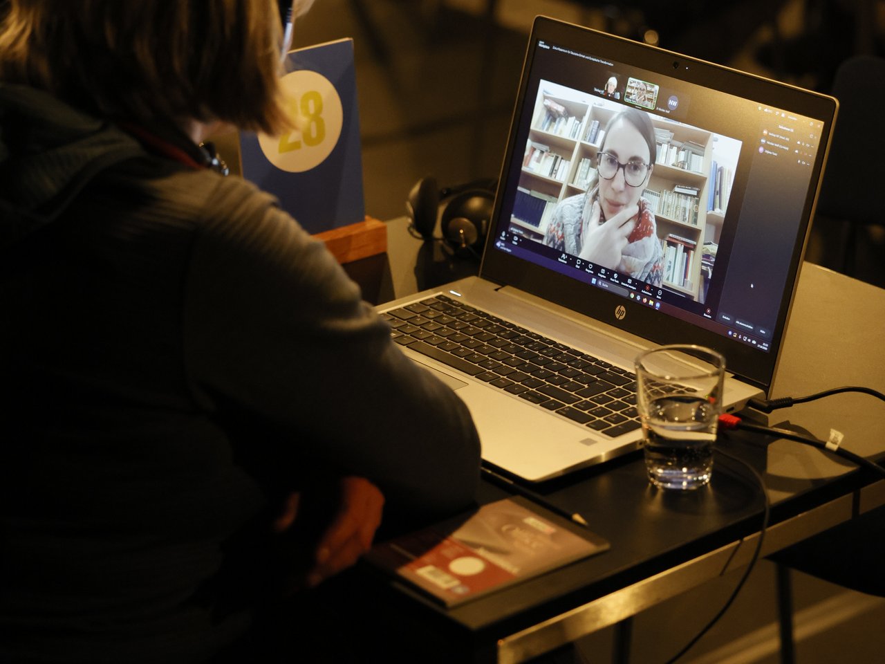 Eine Besucherin mit kurzen blonden Haaren und Kopfhörern sitzt beim Markt für nützliches Wissen und Nicht-Wissen im neuen theater in Halle (Saale) an Tisch Nummer 28. Über einen Laptop führt sie ein Videogespräch mit einer zugeschalteten Expertin, die vor einem Bücherregal sitzt. Auf dem Tisch stehen ein Wasserglas und liegen Informationsmaterialien. Das Gespräch findet digital statt, während ringsum weitere Gespräche in der Arena laufen.