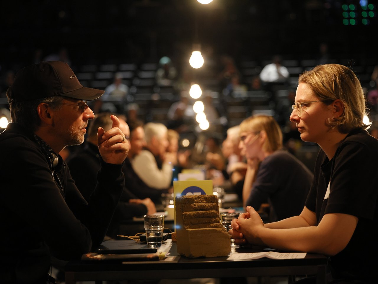 Zwei Personen sitzen sich beim Markt für nützliches Wissen und Nicht-Wissen im neuen theater in Halle (Saale) an einem Tisch gegenüber. Ein Mann mit schwarzer Baseballkappe, Brille und Kopfhörern um den Hals spricht und gestikuliert mit einer Hand. Ihm gegenüber hört eine Frau mit kurzen blonden Haaren und Brille aufmerksam zu. Zwischen ihnen steht ein Modell aus mehreren übereinanderliegenden Erd- oder Materialschichten. Im Hintergrund finden weitere Gespräche in der Arena statt.