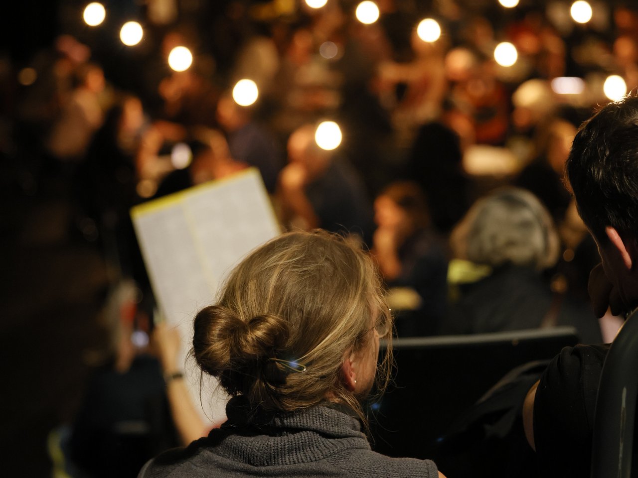 Blick von den ansteigenden Zuschauerreihen auf die Arena beim Markt für nützliches Wissen und Nicht-Wissen im neuen theater in Halle (Saale). Im Vordergrund sitzt eine Frau mit zum Dutt gebundenen blonden Haaren und blickt auf das Geschehen. Im unscharfen Hintergrund finden zahlreiche Einzelgespräche statt. Die vielen hängenden Glühbirnen tauchen den abgedunkelten Veranstaltungsraum in warmes Licht.