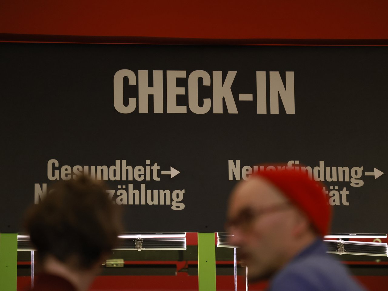 Große weiße Buchstaben mit der Aufschrift „CHECK-IN“ kennzeichnen den Empfangsbereich beim Markt für nützliches Wissen und Nicht-Wissen im neuen theater in Halle (Saale). Darunter weisen Schilder auf die Themenbereiche „Gesundheit – Nacherzählung“ und „Neuerfindung – Stabilität“ hin. Im unscharfen Vordergrund gehen zwei Besucher vorbei, einer trägt eine auffällige rote Mütze.