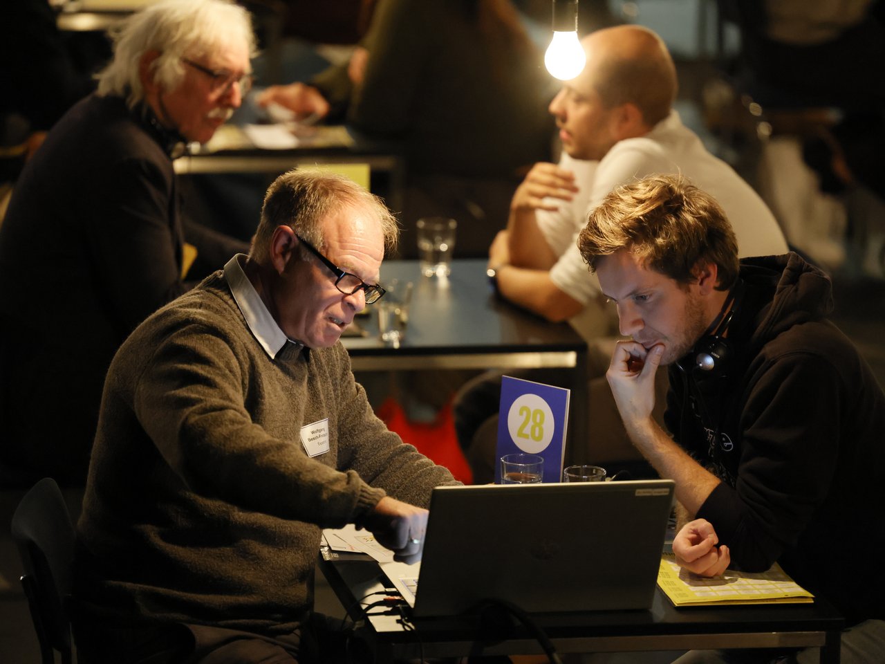 Zwei Männer sitzen sich beim Markt für nützliches Wissen und Nicht-Wissen im neuen theater in Halle (Saale) an Tisch Nummer 28 gegenüber. Ein älterer Mann mit Brille und grauem Pullover zeigt seinem jüngeren Gesprächspartner etwas auf einem Laptop. Dieser trägt einen schwarzen Kapuzenpullover und Kopfhörer um den Hals und betrachtet konzentriert den Bildschirm. Im Hintergrund finden weitere Einzelgespräche statt.