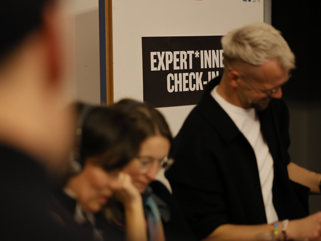 Mehrere Personen stehen beim Expert:innen-Check-in des Marktes für nützliches Wissen und Nicht-Wissen im neuen theater in Halle (Saale). Im Vordergrund sind zwei Frauen zu sehen, die sich über Unterlagen beugen. Rechts steht ein Mann mit kurzen grauen Haaren, Brille und schwarzer Jacke. Im Hintergrund hängt ein schwarzes Schild mit der weißen Aufschrift „EXPERT*INNEN CHECK-IN“.