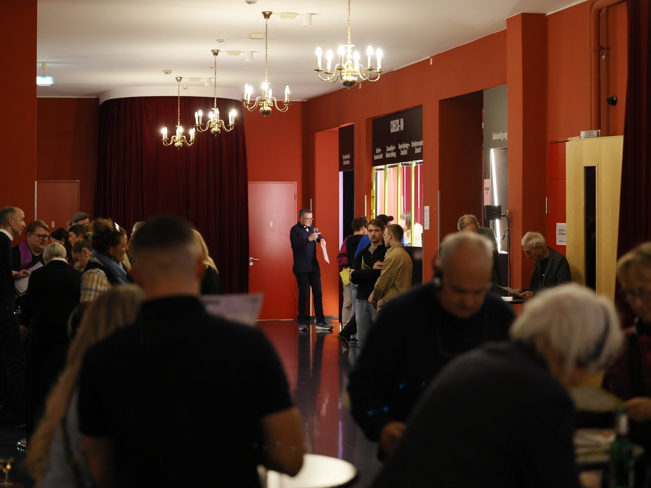 Blick in das gut besuchte Foyer beim Markt für nützliches Wissen und Nicht-Wissen im neuen theater in Halle (Saale). Zahlreiche Besucher:innen stehen in kleinen Gruppen zusammen, unterhalten sich oder lesen das Veranstaltungsprogramm. Im Hintergrund ist der Check-in-Tresen zu sehen. Rote Wände, dunkle Vorhänge und mehrere Kronleuchter prägen den Raum.