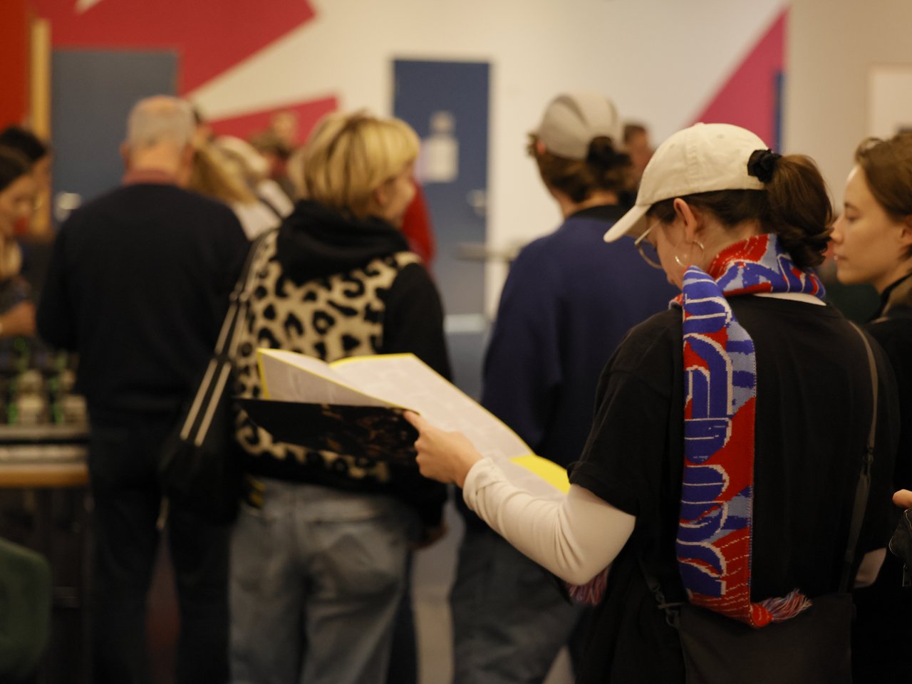 Eine Besucherin mit weißer Baseballkappe und rot-blauem Schal liest aufmerksam die aufgefaltete Enzyklopädie beim Markt für nützliches Wissen und Nicht-Wissen im neuen theater in Halle (Saale). Im Hintergrund stehen weitere Besucher:innen im belebten Foyer, einige unterhalten sich, andere informieren sich über das Angebot.
