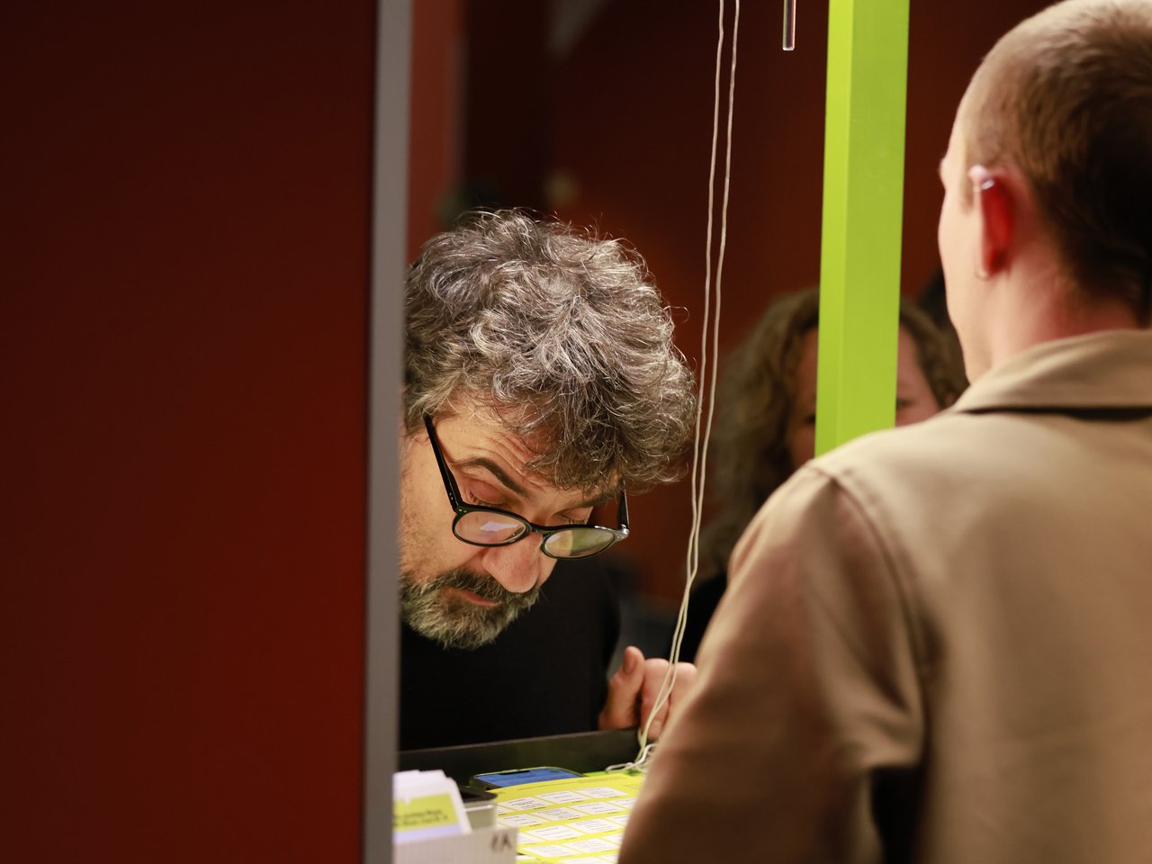 Ein Mann mit grauen Locken, Bart und Brille beugt sich über den Check-in-Tresen beim Markt für nützliches Wissen und Nicht-Wissen im neuen theater in Halle (Saale). Er betrachtet die ausliegenden Unterlagen, während ihm ein Mitarbeiter in beigefarbener Jacke gegenübersteht.