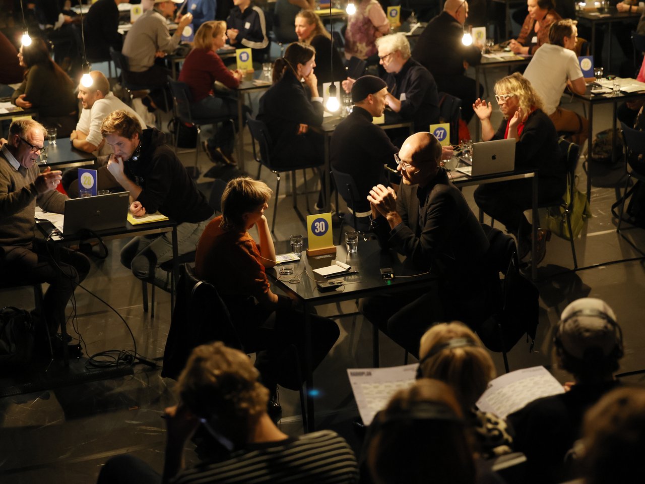 Blick von oben auf die gut besuchte Gesprächsarena beim Markt für nützliches Wissen und Nicht-Wissen im neuen theater in Halle (Saale). An zahlreichen nummerierten Tischen sitzen sich Expert:innen und Besucher:innen gegenüber und tauschen sich aus. Einige gestikulieren lebhaft, andere hören aufmerksam zu. Auf den Tischen stehen Laptops, Wassergläser und Gesprächsunterlagen. Im Vordergrund verfolgen weitere Gäste die Gespräche über Kopfhörer. Hängende Glühbirnen beleuchten den abgedunkelten Raum.