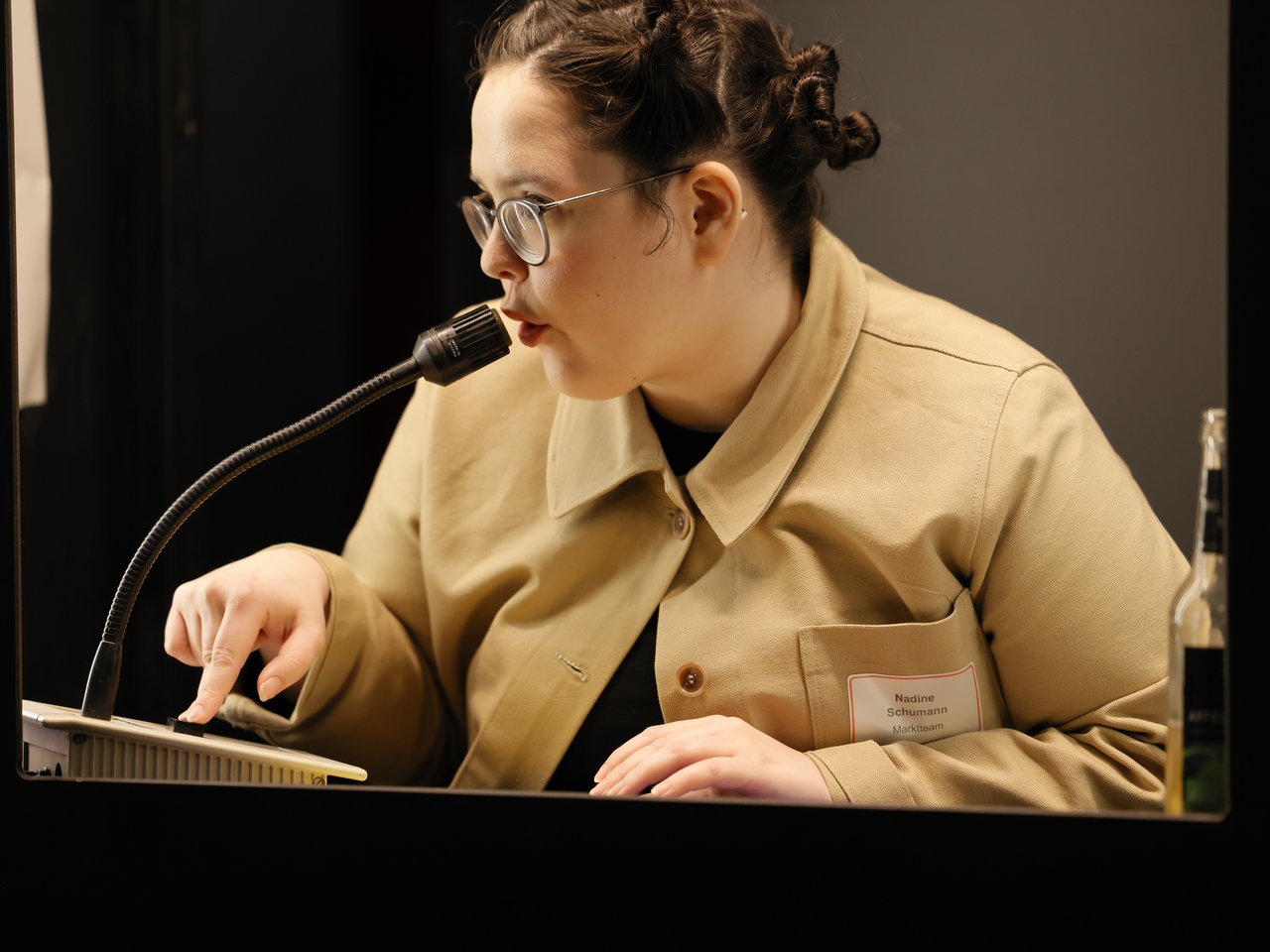 Nadine Schumann vom Marktteam spricht beim Markt für nützliches Wissen und Nicht-Wissen im neuen theater in Halle (Saale) in ein Mikrofon. Sie trägt eine beigefarbene Jacke und eine Brille, ihre Haare sind zu kleinen Dutts hochgesteckt. Während sie spricht, bedient sie mit einem Finger ein Gerät vor sich.