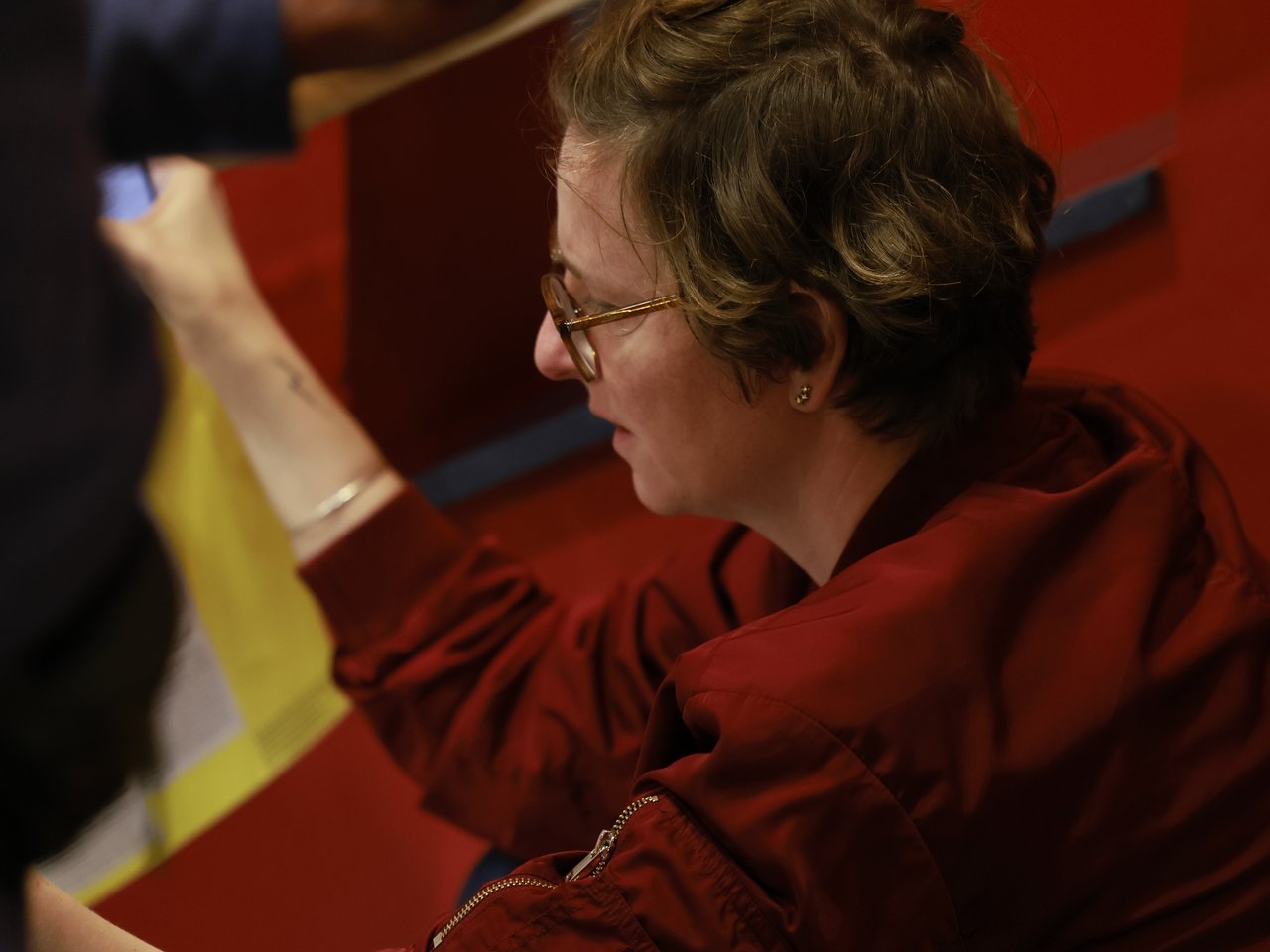 Eine Frau mit kurzen braunen Haaren, Brille und roter Jacke sitzt beim Markt für nützliches Wissen und Nicht-Wissen im neuen theater in Halle (Saale). Sie betrachtet eine Enzyklopädie und spricht mit einer Person, die im Vordergrund ein Smartphone und ebenfalls ein Programm in den Händen hält.