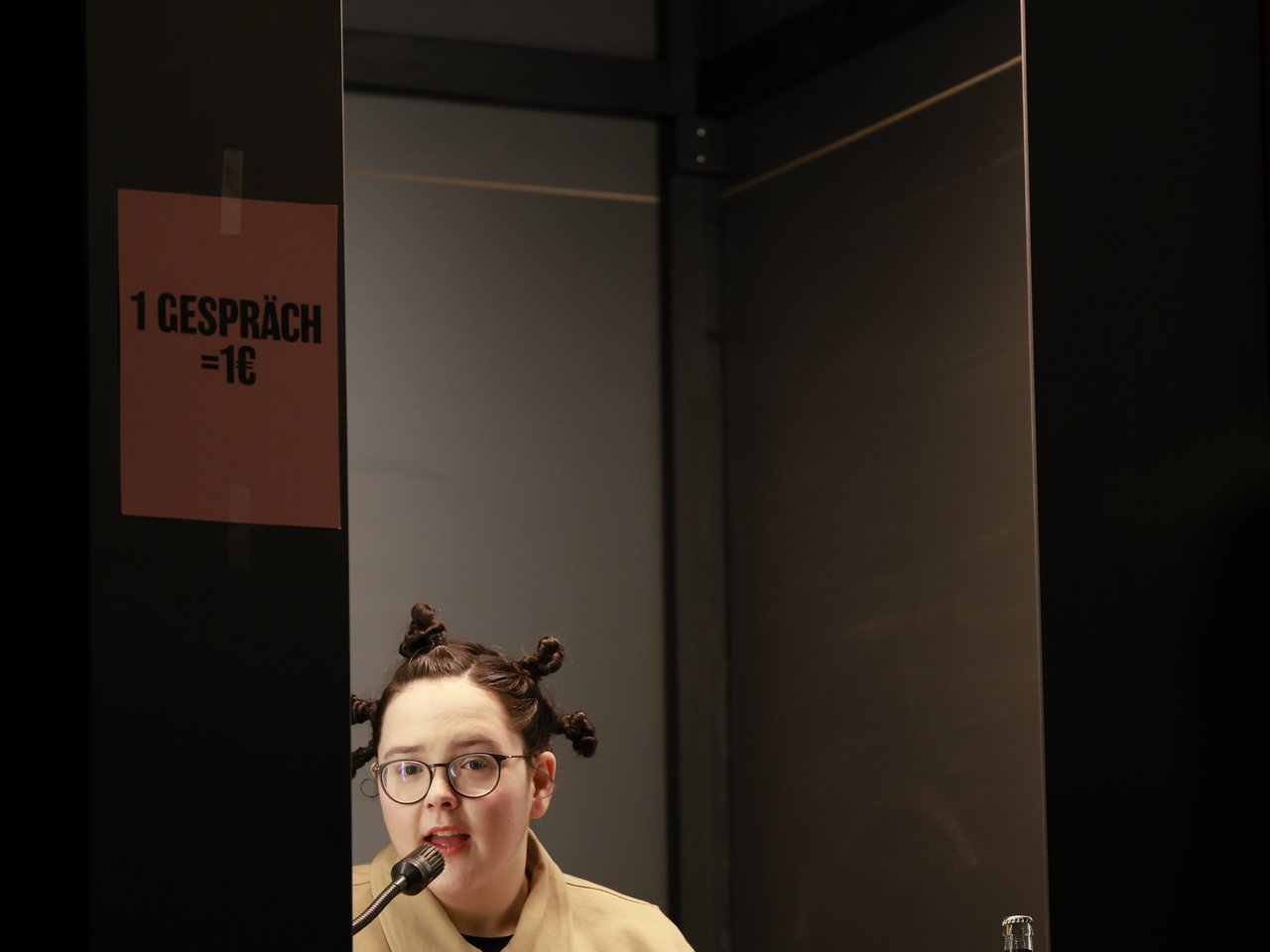 Eine Mitarbeiterin mit Brille und hochgesteckten Haaren spricht in ein Mikrofon am Ankündigungsschalter beim Markt für nützliches Wissen und Nicht-Wissen im neuen theater in Halle (Saale). Über dem Schalter steht „Ankündigungen“, daneben hängt ein Schild mit der Aufschrift „1 Gespräch = 1 €“.