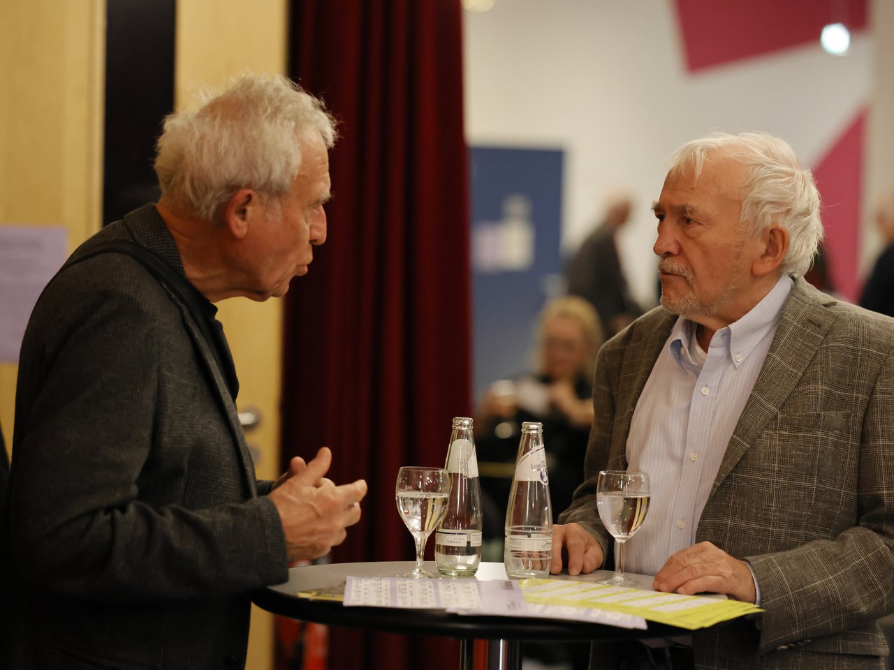 Zwei ältere Männer mit weißen Haaren stehen beim Markt für nützliches Wissen und Nicht-Wissen im neuen theater in Halle (Saale) an einem Stehtisch und unterhalten sich. Der Mann links trägt eine dunkle Jacke und gestikuliert mit den Händen, während sein Gegenüber in einem grauen Sakko aufmerksam zuhört. Auf dem Tisch stehen zwei Wasserflaschen und zwei Gläser, daneben liegen Enzyklopädien.