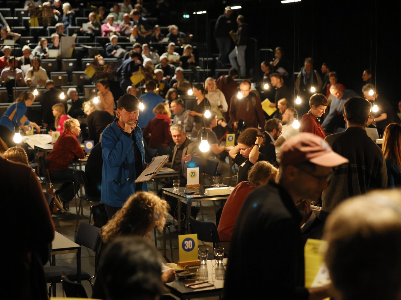 Blick in die gut besuchte Arena beim Markt für nützliches Wissen und Nicht-Wissen im neuen theater in Halle (Saale). Zahlreiche Besucher:innen und Expert:innen verteilen sich auf kleine, nummerierte Gesprächstische, während weitere Gäste auf den ansteigenden Sitzreihen Platz nehmen. Im Vordergrund steht ein Mann mit blauer Jacke und schwarzer Kappe, der ein Klemmbrett in der Hand hält. Hängende Glühbirnen beleuchten den lebendigen Veranstaltungsraum.