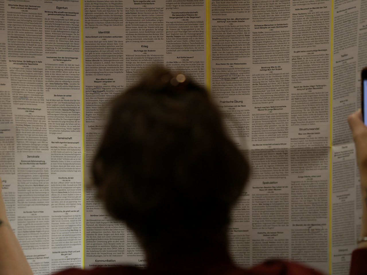 Eine Besucherin betrachtet die großformatige, aufgefaltete Enzyklopädie des Marktes für nützliches Wissen und Nicht-Wissen im neuen theater in Halle (Saale). Sie enthält zahlreiche Gesprächsangebote, die in mehreren Spalten nach Themen geordnet sind. Im Vordergrund ist die Besucherin von hinten zu sehen, rechts hält sie ein Smartphone in der Hand.