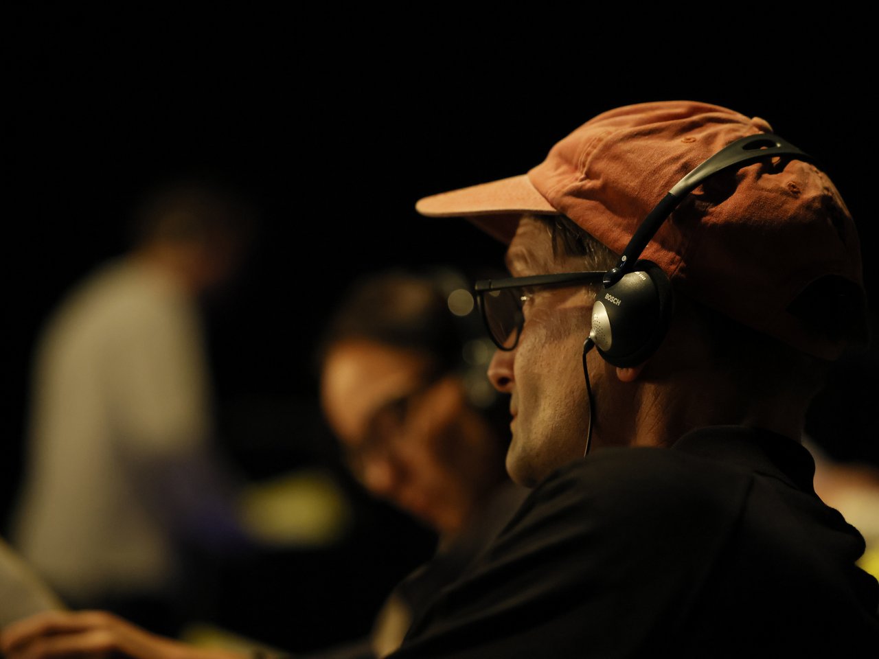 Ein Besucher mit orangefarbener Baseballkappe, Brille und Kopfhörern sitzt beim Markt für nützliches Wissen und Nicht-Wissen im neuen theater in Halle (Saale) auf den Zuschauerplätzen. Er blickt in Richtung der Gesprächsarena und hält eine Enzyklopädie in der Hand. Im unscharfen Hintergrund sind weitere Besucher:innen zu sehen. Über die Kopfhörer können die Gäste ausgewählte Einzelgespräche live mitverfolgen.