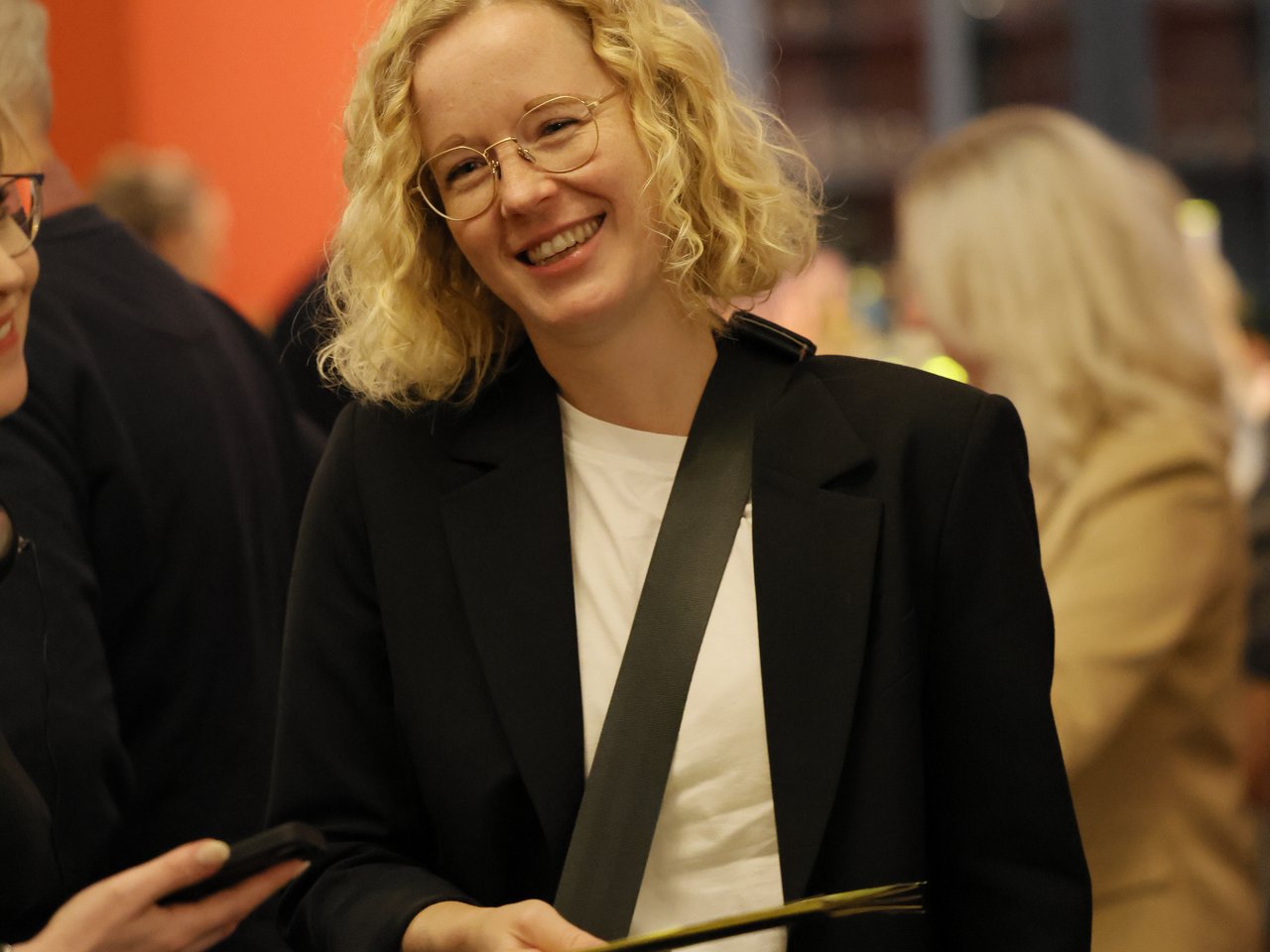 Dr. Uta Bretschneider, Direktorin des Zukunftszentrums, steht lächelnd beim Markt für nützliches Wissen und Nicht-Wissen im neuen theater in Halle (Saale). Sie trägt einen schwarzen Blazer über einem weißen Oberteil, eine Brille und eine Umhängetasche. Im Hintergrund sind weitere Gäste im Foyer zu sehen.